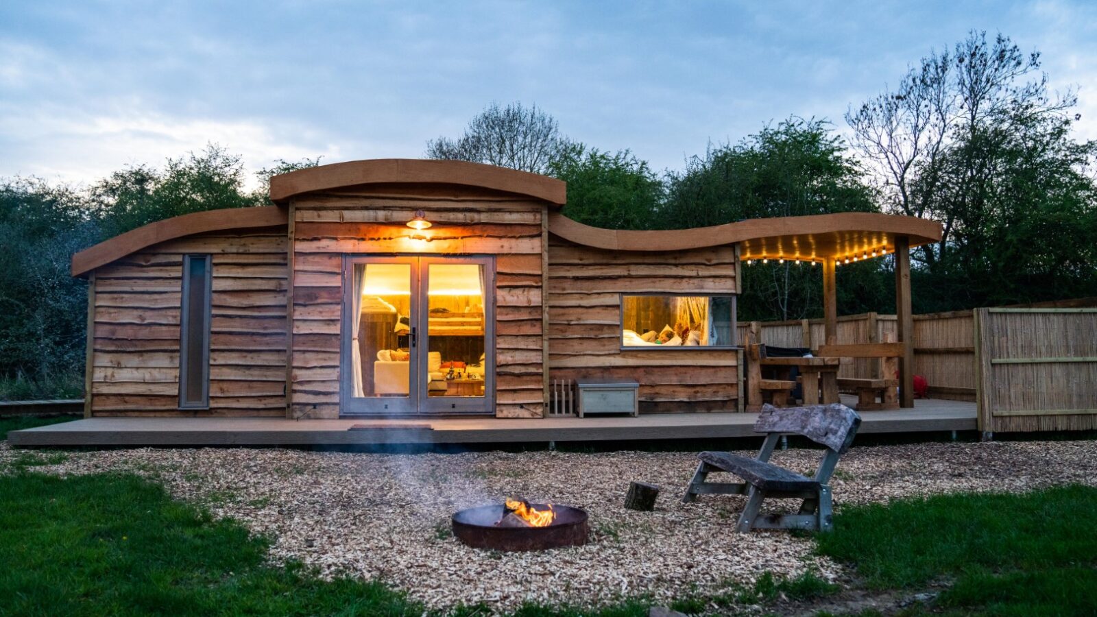 A modern wooden kabina with large windows and a lit interior exudes warmth. A campfire burns in front, surrounded by lush grass and trees, reminiscent of the tranquil charm found at Baxby Manor.