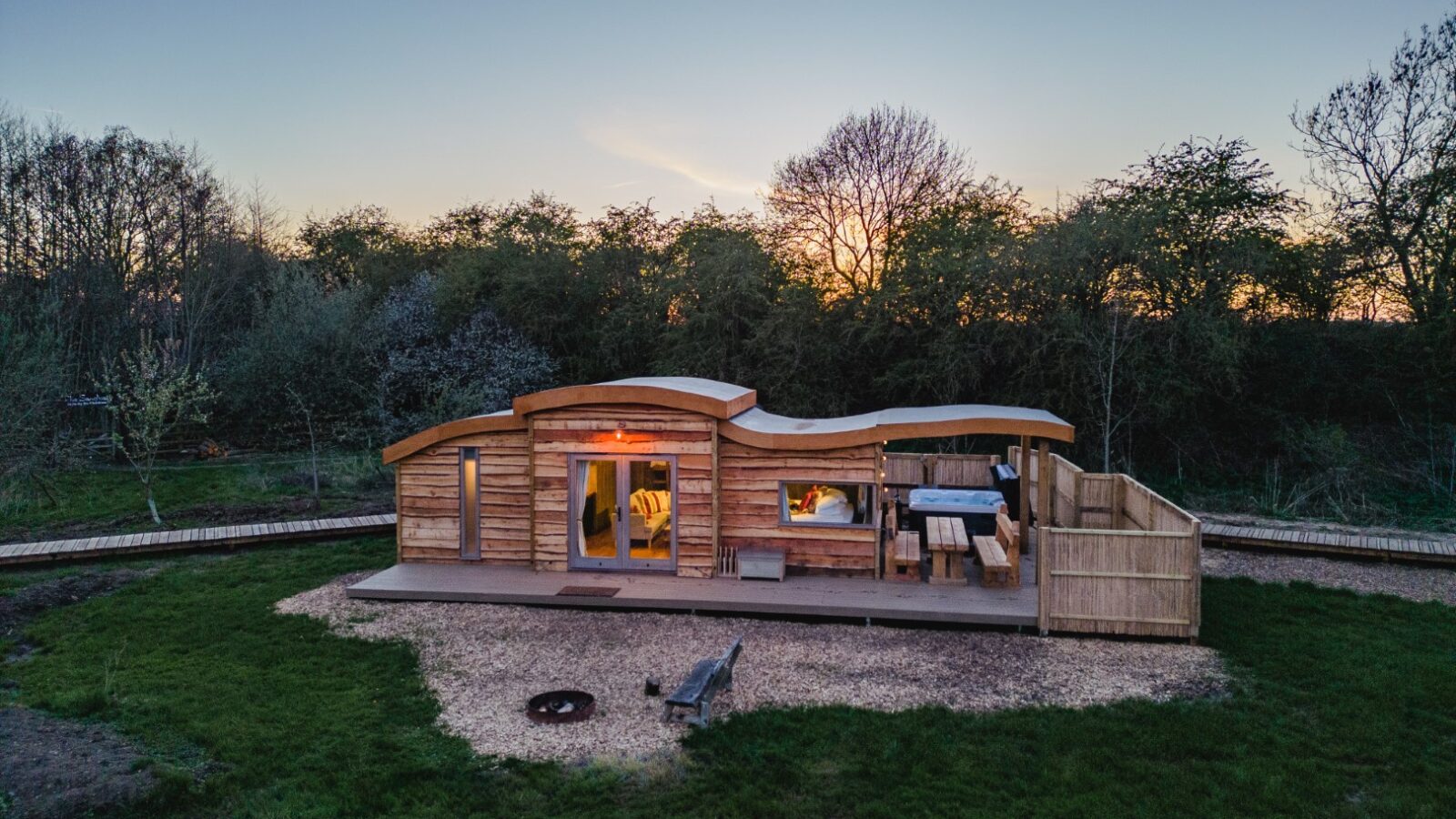 A small wooden cabin with a curved roof and outdoor seating area sits in a grassy clearing surrounded by trees at dusk, reminiscent of the charming retreats found at Baxby Manor.