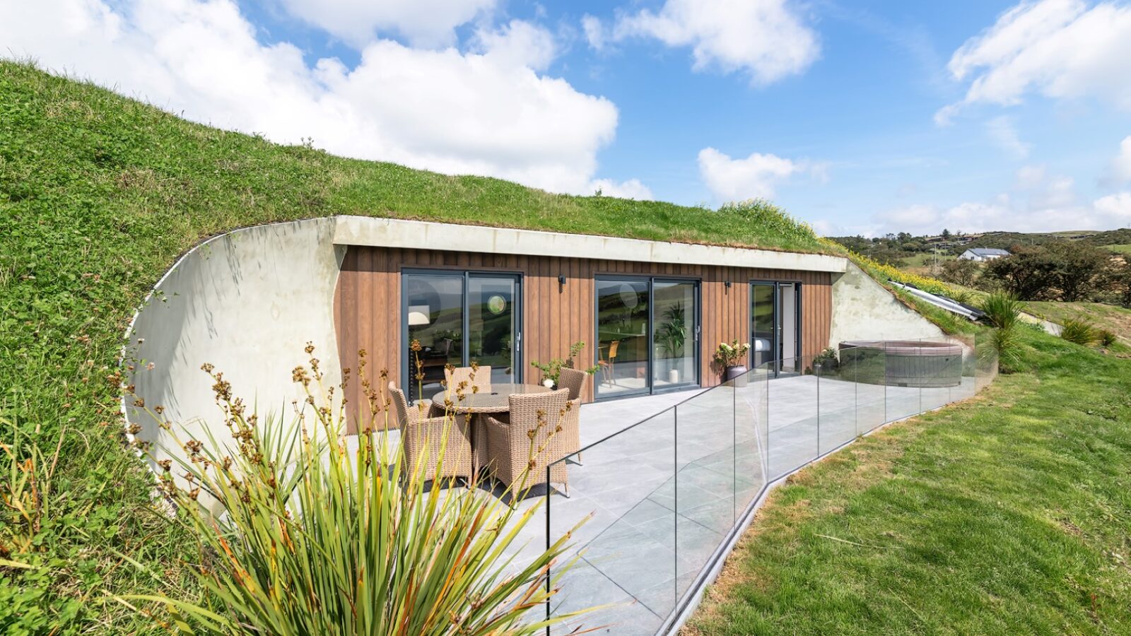 Nestled in the serene landscape of Willow Hill, this modern earth-sheltered house features a striking glass facade and a charming patio, all embraced by lush greenery under a partly cloudy sky.