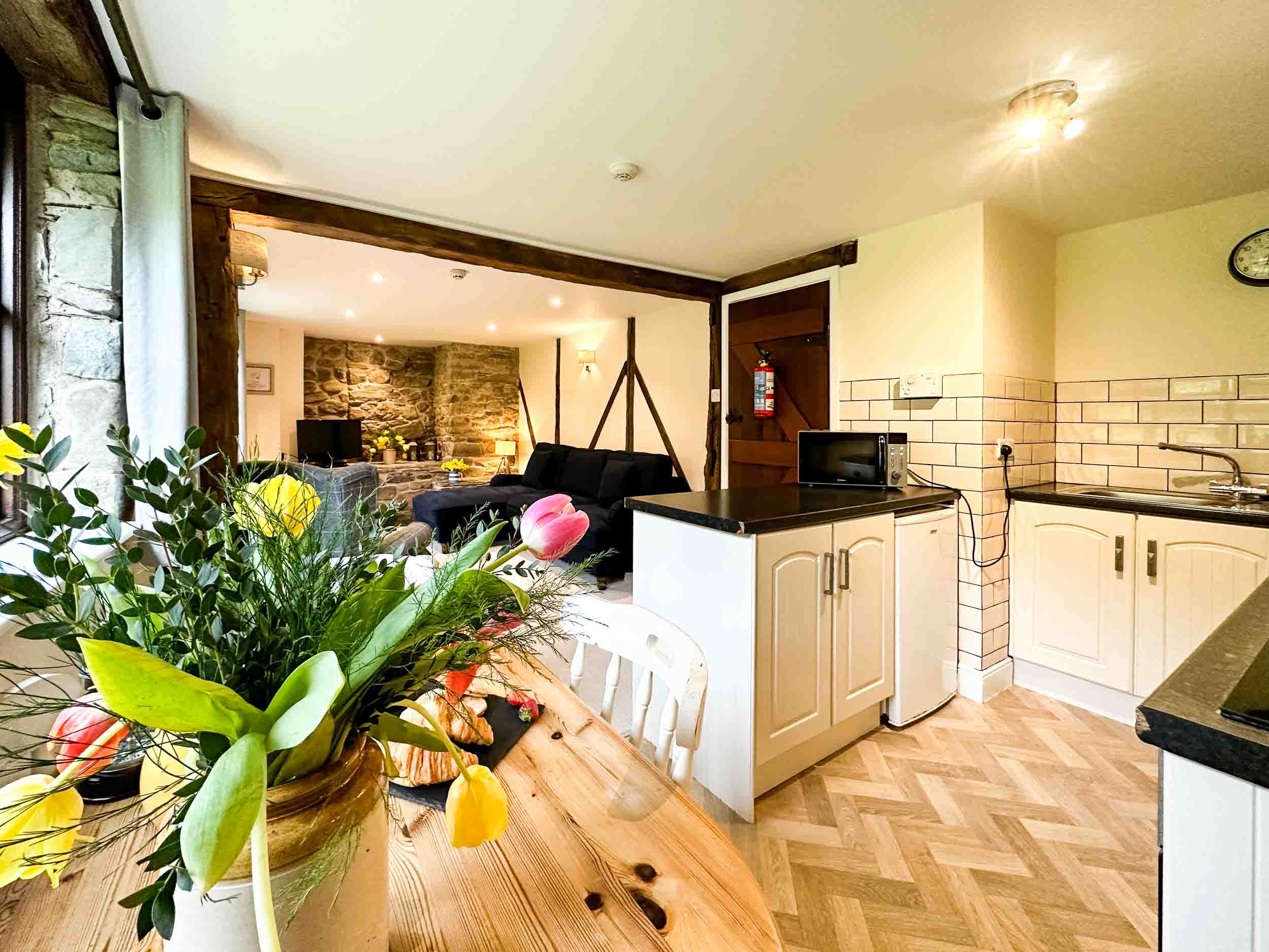 A cosy kitchen with light wood cabinetry and a black countertop, which includes a sink and microwave. The adjacent dining table has a vase with colourful flowers. The room opens into a living area with a black sofa and stone accent wall, visible through a wooden beam partition – perfect for a Family Getaway UK.