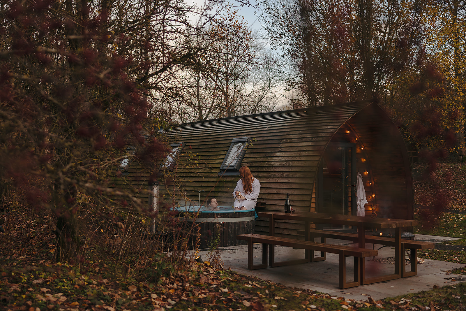 A person in a hot tub next to a wooden cabin is surrounded by trees and autumn leaves, embodying the essence of winter glamping in the UK. A picnic table nearby completes this serene escape.