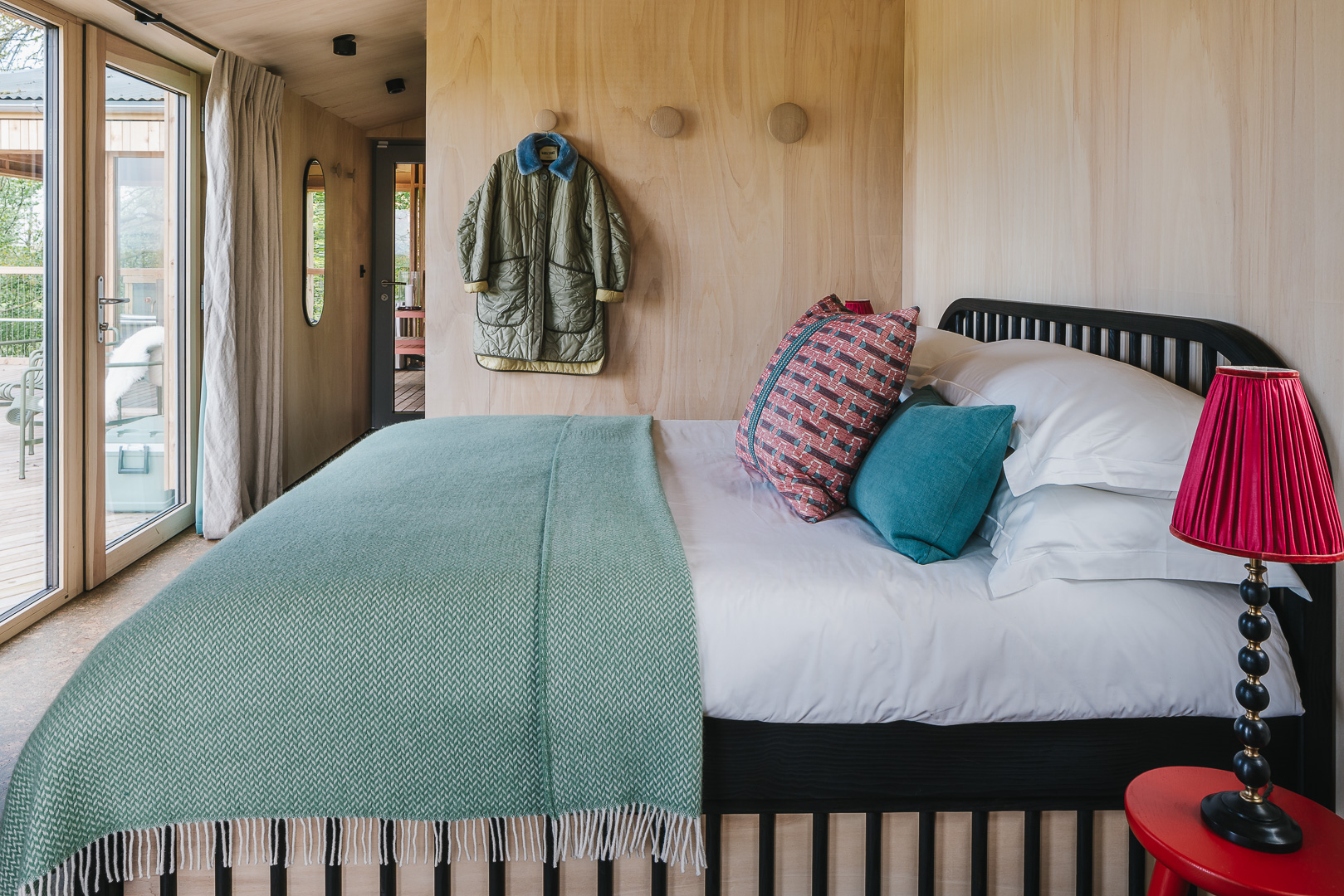 A cozy bedroom showcases a neatly made bed with colorful pillows, a red lamp, and a coat on the wall. To enhance your holiday let website photos, make sure to highlight features like the sliding glass door leading outside, ensuring prospective guests feel welcomed and intrigued.