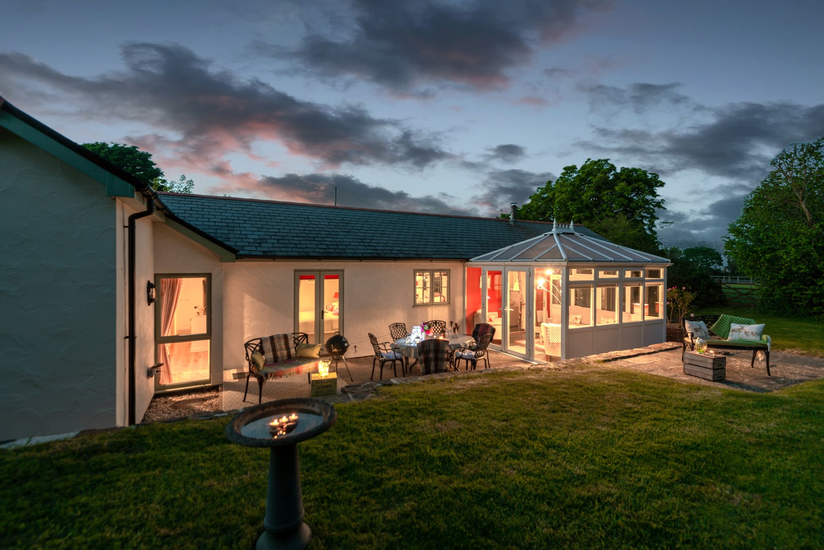 This single-story house, perfect for holiday let owners seeking inspiration to improve their property photos, boasts a glass-roofed conservatory. Lit beautifully at dusk, it features inviting outdoor seating and a lush garden with trees—a picturesque setting that speaks for itself.