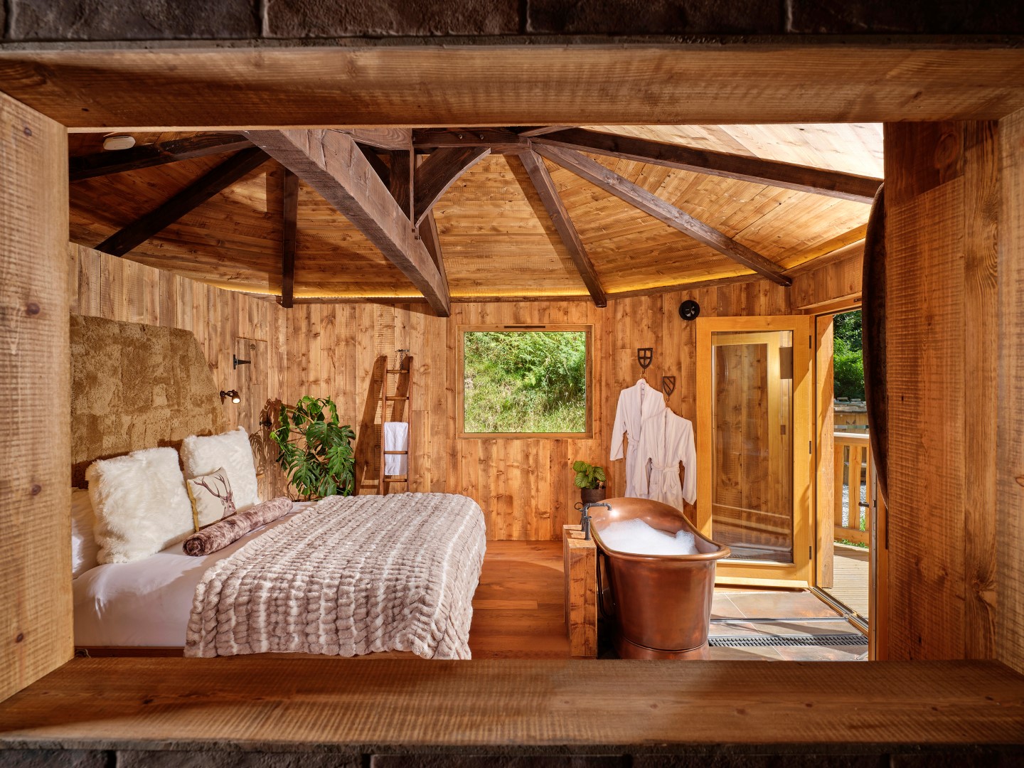 A cozy wooden cabin interior features a comfortable bed, lush plants, and a copper bathtub. Robes hang nearby while a large window frames the serene greenery outside—perfect for showcasing on your website. Remember these 3 top tips for holiday let owners to enhance your photos: lighting, angles, and decluttered spaces.