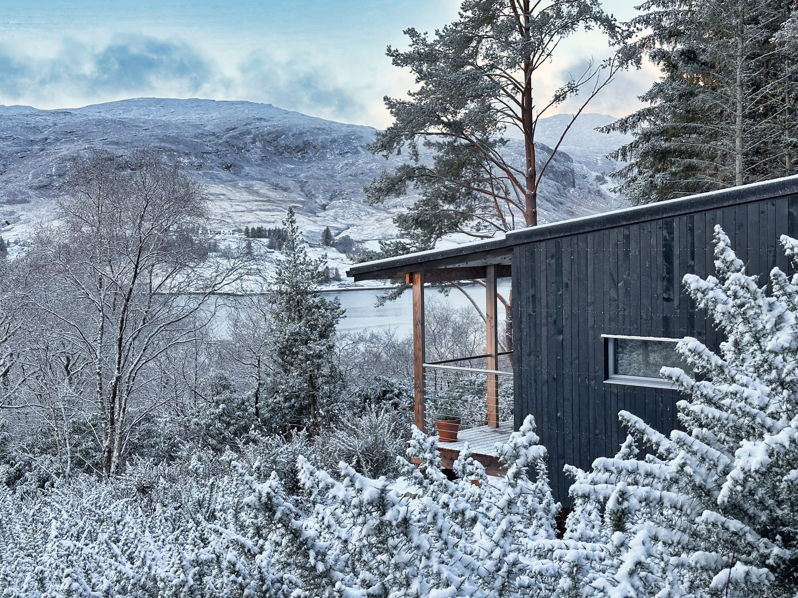 A modern cabin with glass windows offers an exquisite winter glamping experience, surrounded by snow-covered trees and hilly landscapes under a partly cloudy sky in the UK.