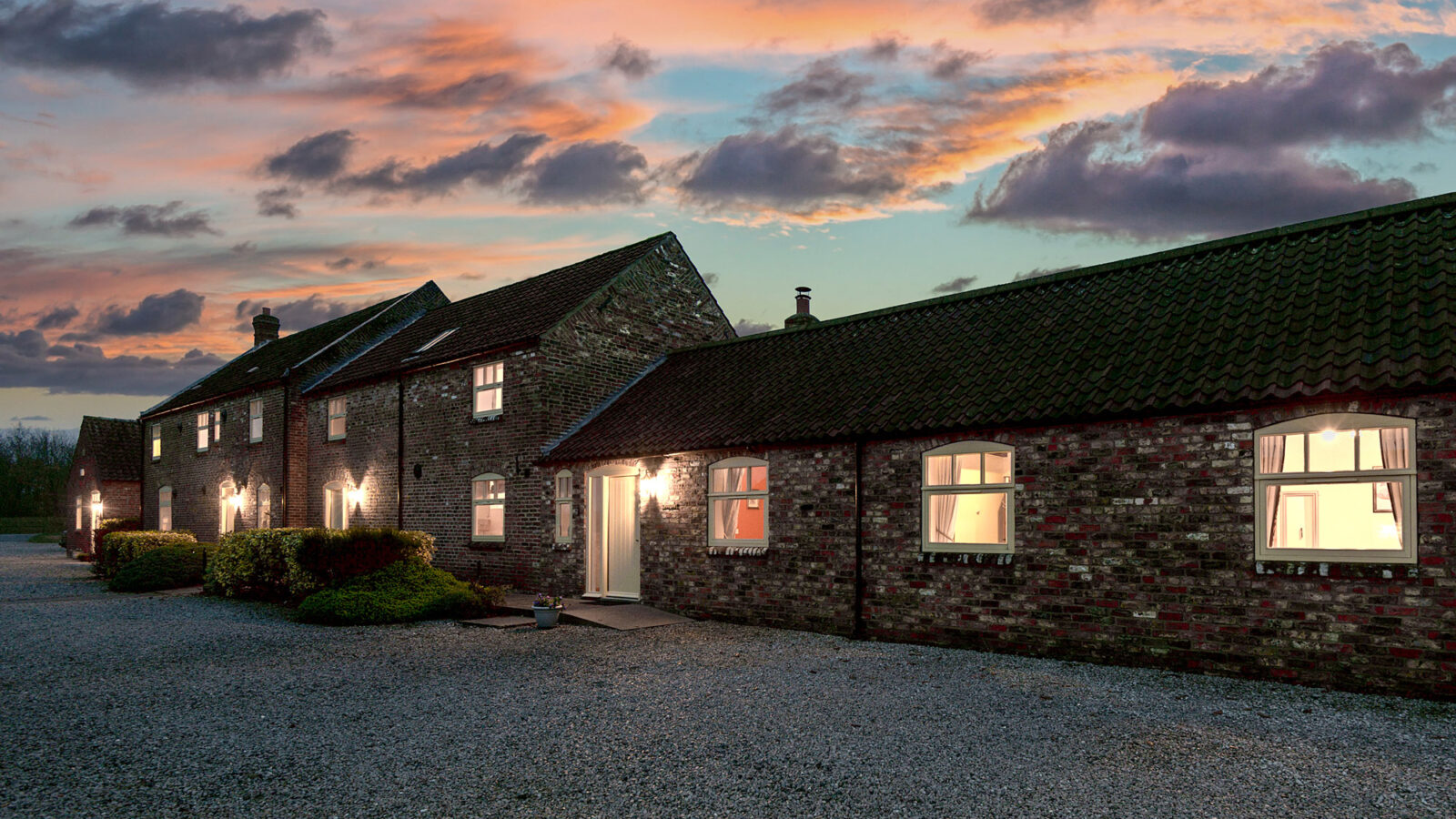 A charming brick house with illuminated windows and a gravel driveway at sunset evokes the cozy ambiance of Broadgate Farm Cottages, nestled under a sky adorned with clouds.