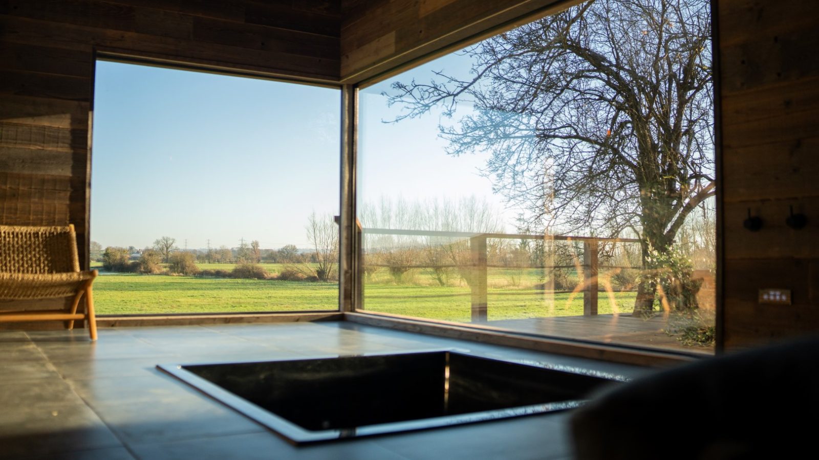 A sunlit room with large windows overlooks a grassy orchard and trees, featuring a recessed tub and a wicker chair.