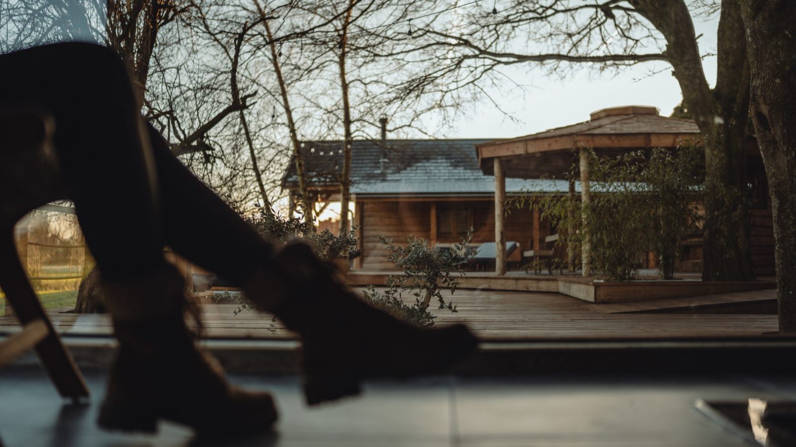 Silhouette of a boot-clad person indoors, gazing through a large window at an orchard and wooden cabin amidst trees.