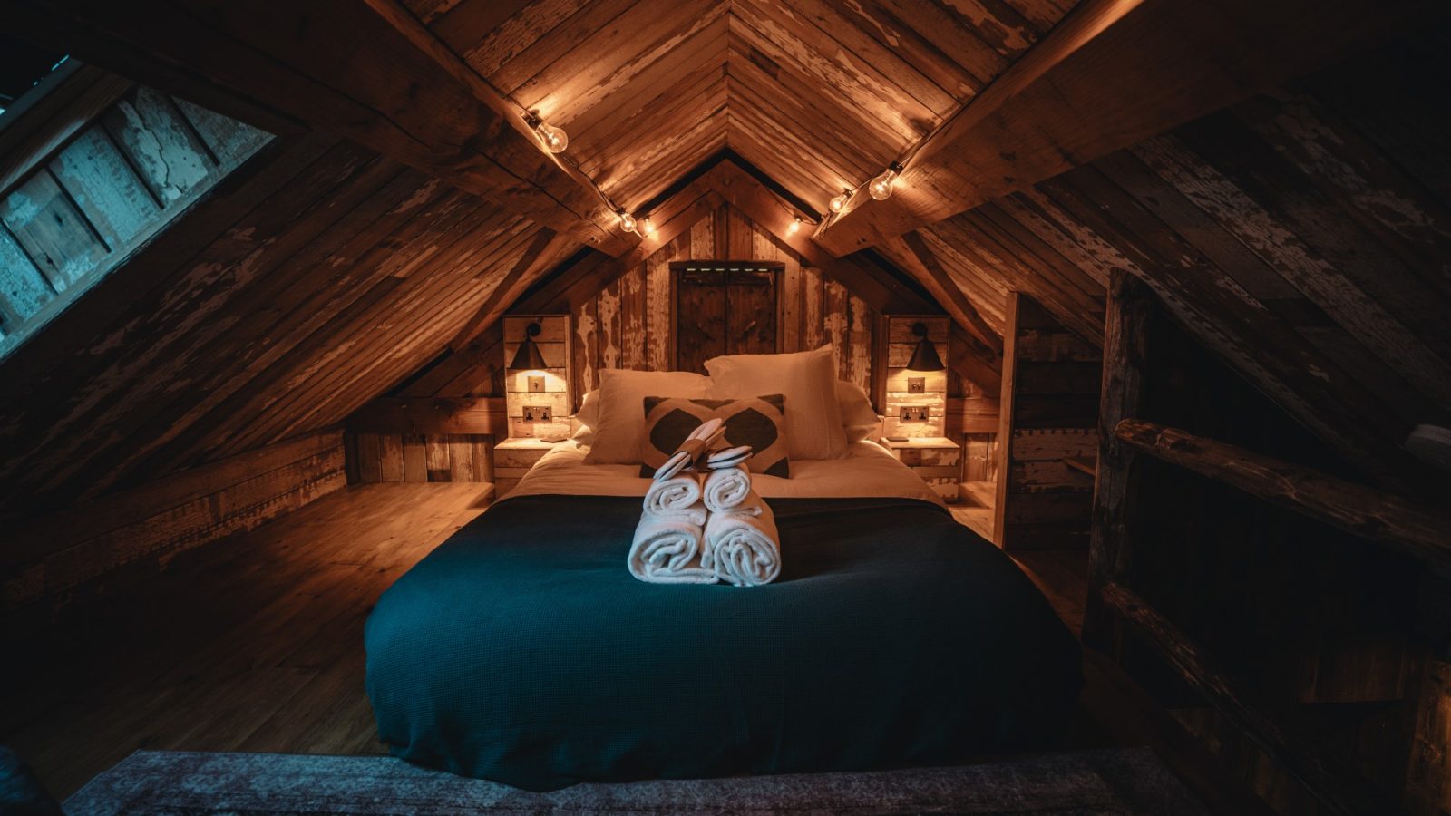 Cozy attic bedroom with wooden walls, bed with teal blanket, and towels folded into swans, warmly lit near Fenny Castle.