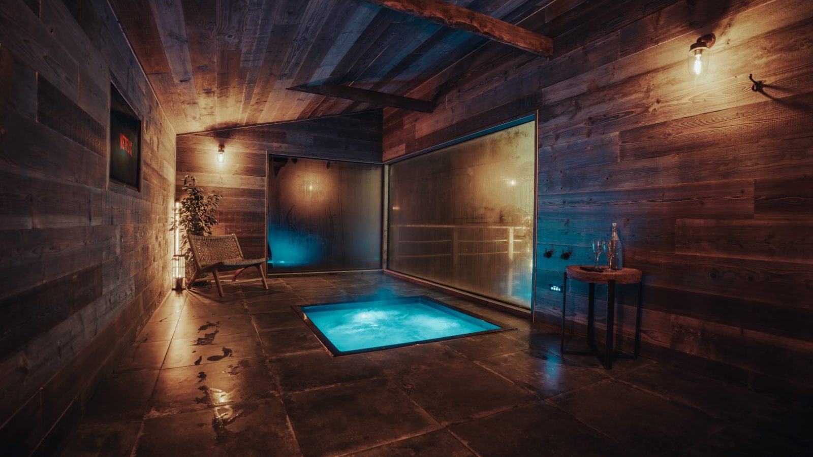 A dimly lit room with wooden walls features a small illuminated pool, a chair, and a side table with glasses and an Orchard bottle.