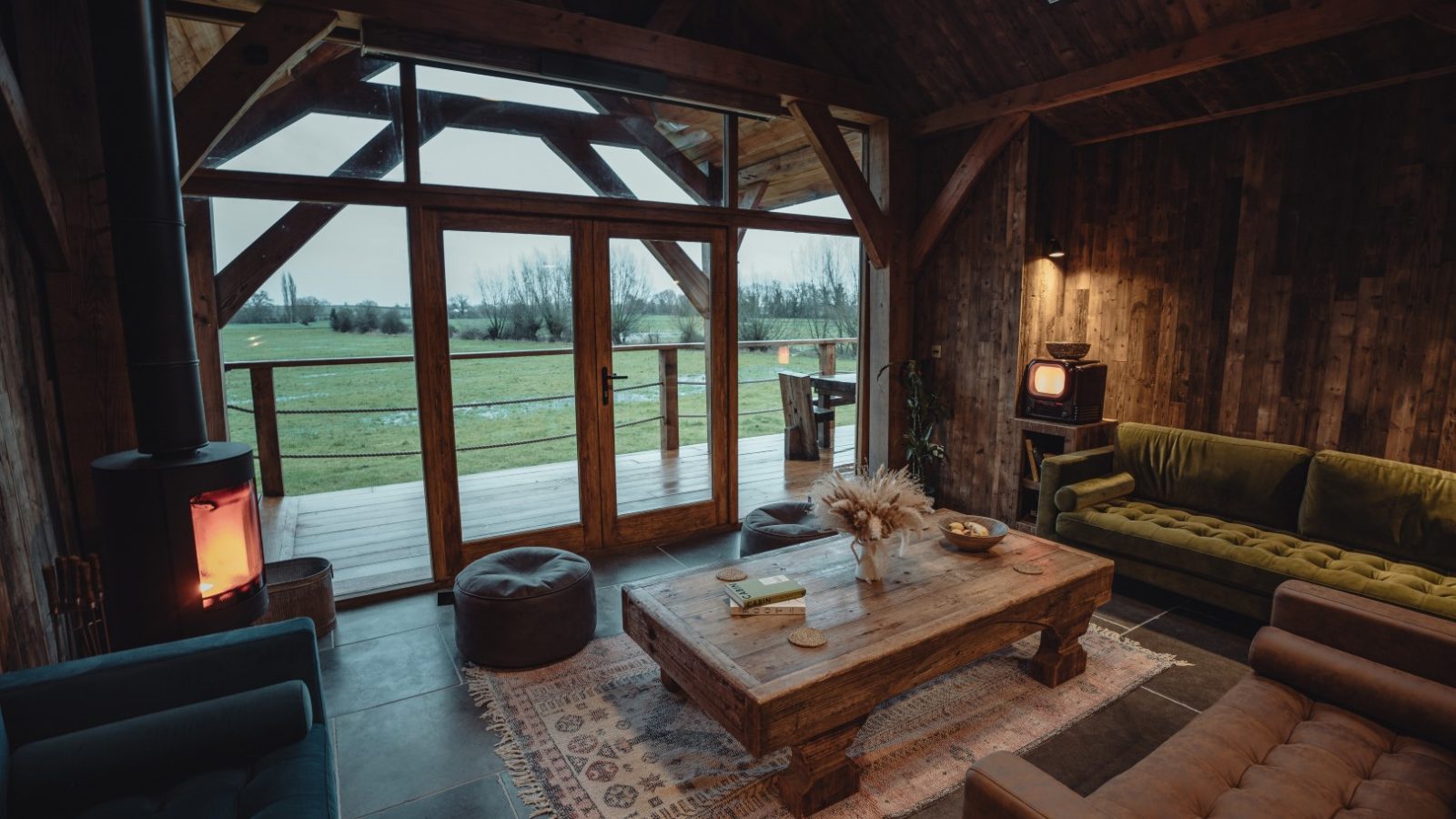 Cozy wooden cabin interior with a large coffee table, sofas, and a fireplace, offering views of the orchard through glass doors.