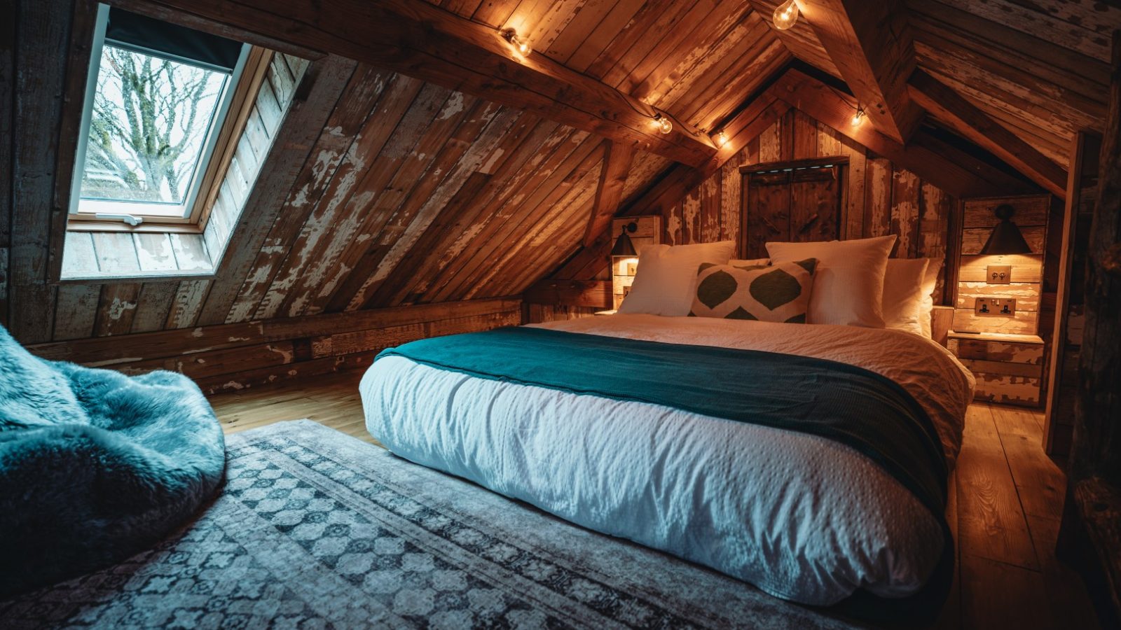 Cozy attic bedroom with wooden walls, double bed, string lights, a skylight, and a patterned rug overlooking Fenny Castle.