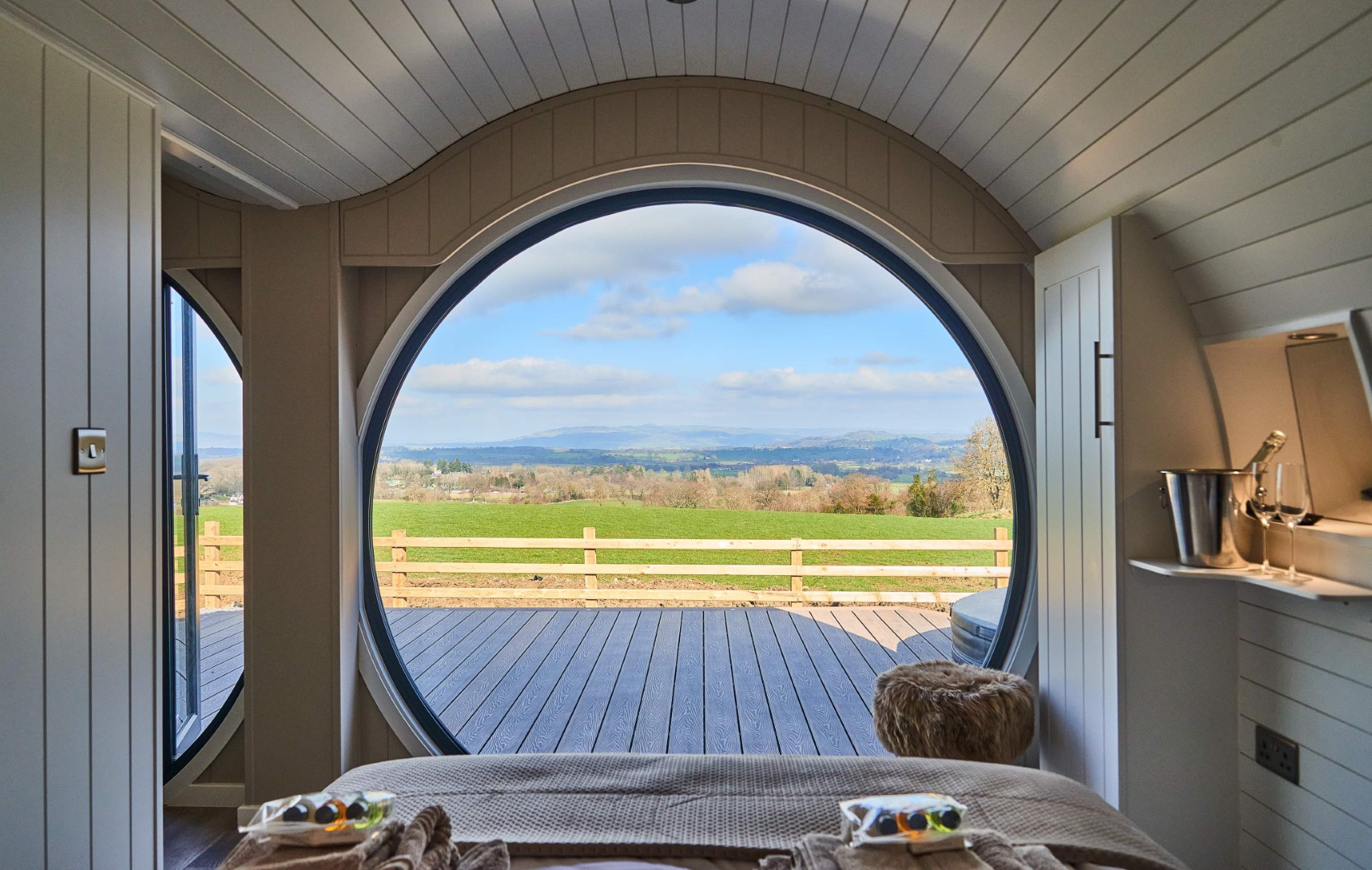 A cosy room with a circular window offers a scenic view of green fields and distant hills, perfect for winter glamping in the UK. The room has light wooden panelling and a plush pouf. The open window space extends to a wooden deck, adding to the serene and inviting ambiance.