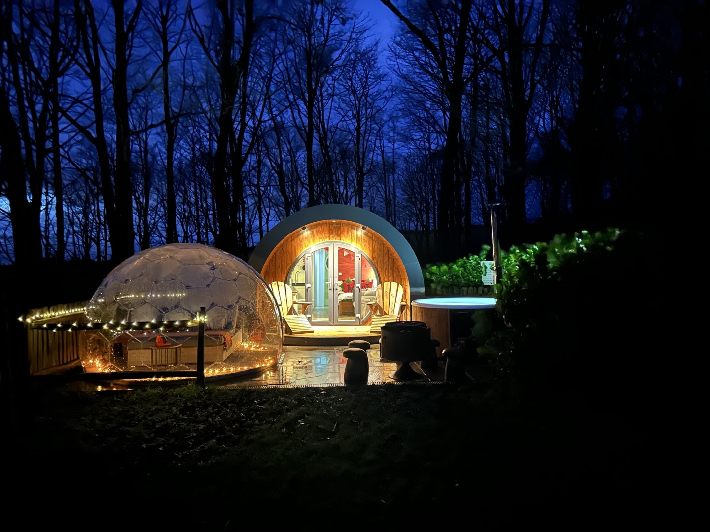 A cosy glamping pod with a geodome on the patio is illuminated at night in a forest setting, perfect for winter glamping in the UK. Warm lights outline the structures and pathways, creating a serene and inviting atmosphere. Fairy lights decorate the area, and an outdoor hot tub is visible in the background.