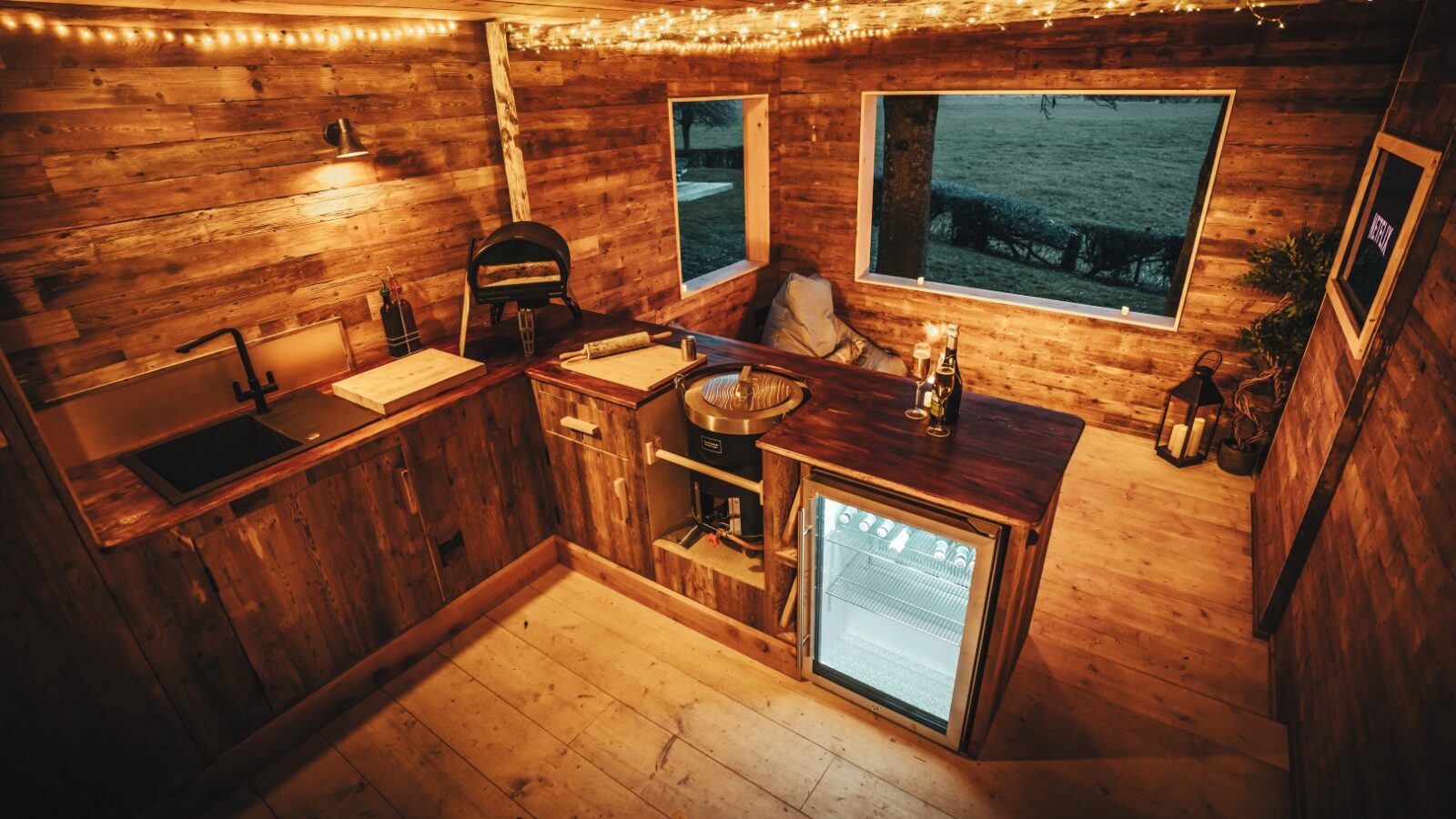 Cozy wooden kitchen interior with a counter, mini-fridge, sink, and string lights. A large window offers a charming view of The Orchard outside.