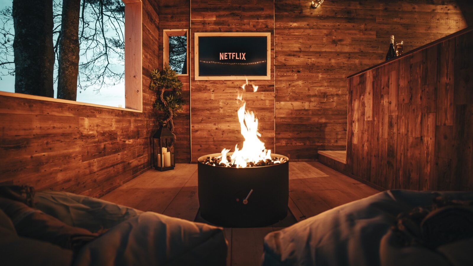 Cozy wooden room with a fire pit, Netflix on TV, and bean bags. Floor-to-ceiling windows provide a stunning view of trees at dusk, reminiscent of an orchard stroll near Fenny Castle.