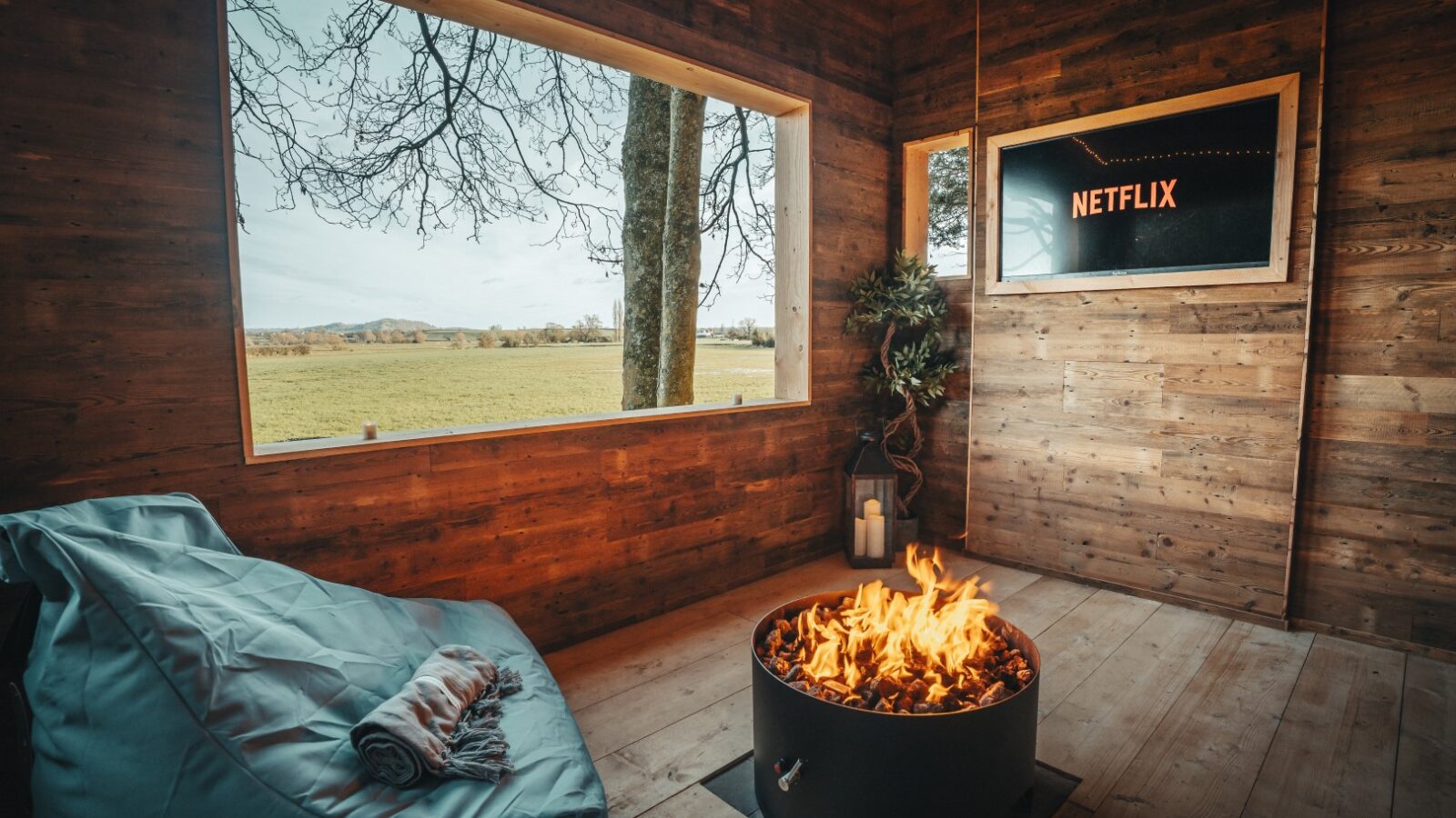 Cozy room with a fire pit, a bean bag chair, and a TV displaying Netflix. The large window offers views of the grassy field and charming Fenny Castle, adding a touch of enchantment to the peaceful setting.