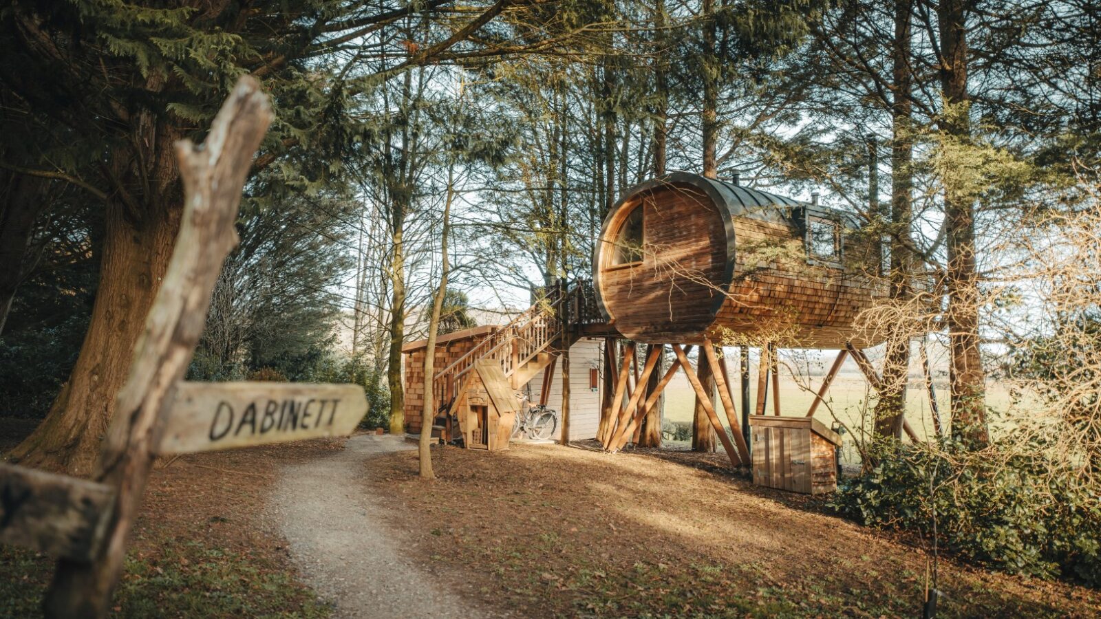 A wooden treehouse with a cylindrical design elevated on stilts among the trees, nestled in an orchard. A pathway leads to a sign that reads 