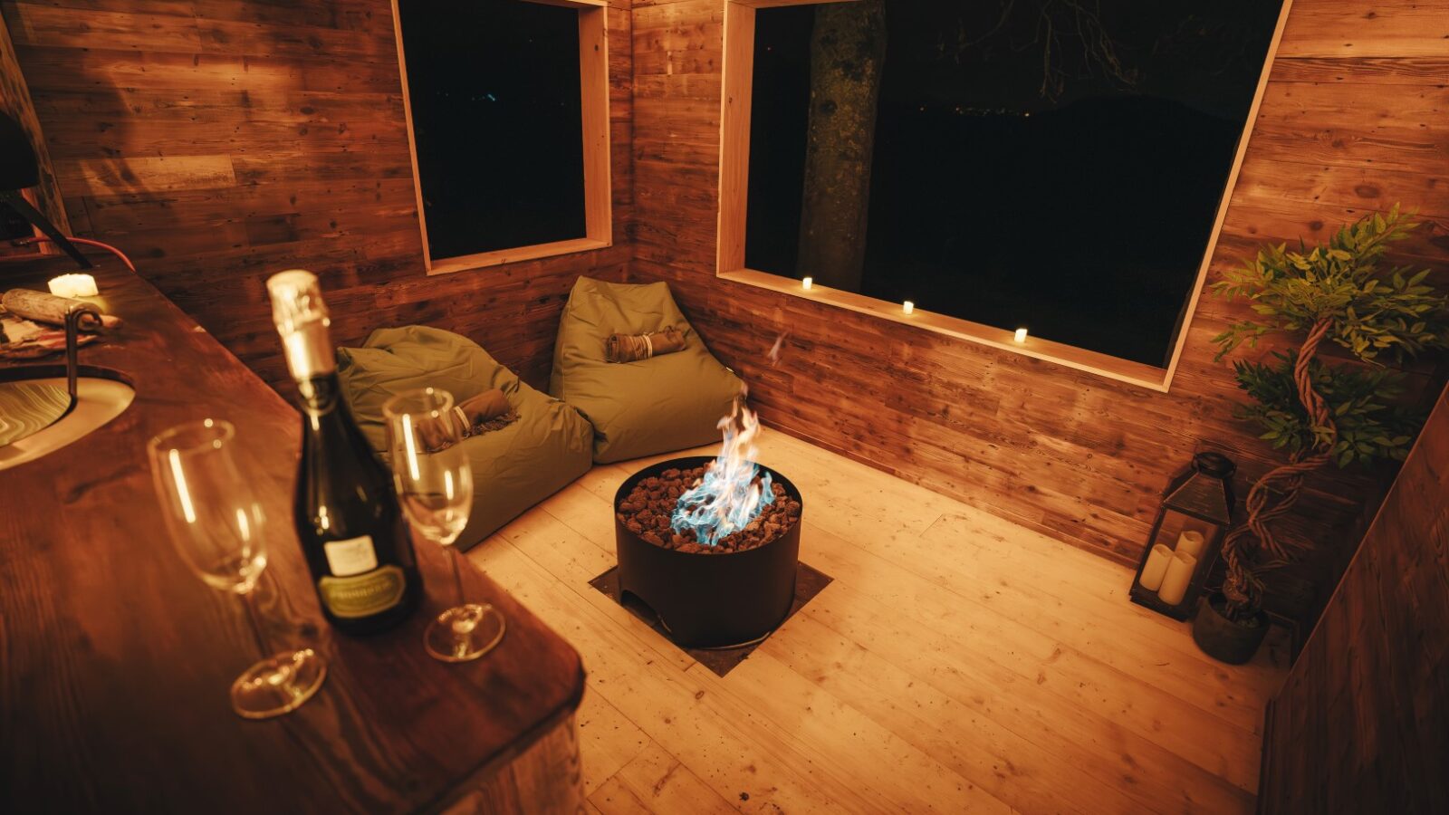 Cozy wooden room with bean bags, a small fire pit, wine glasses, and a bottle of wine on a counter, softly lit by candlelight. The gentle ambiance conjures the charm of Fenny Castle and the serenity found in an orchard.