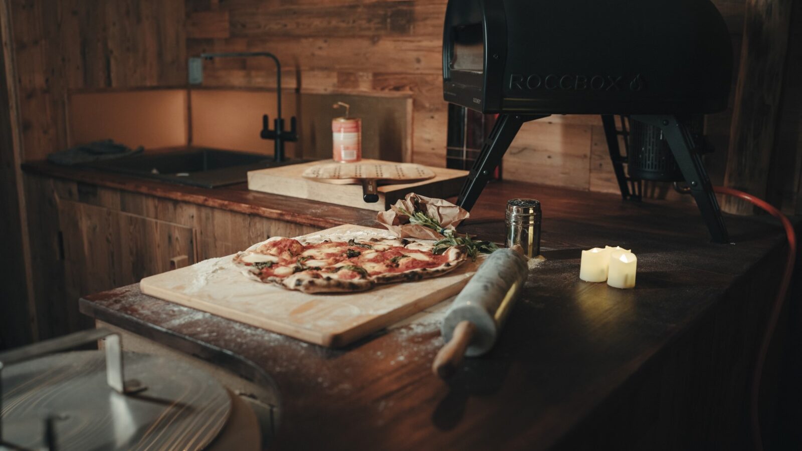 A Fenny Castle wood-fired pizza rests on a wooden board near a Roccbox pizza oven, surrounded by flickering candles and essential kitchen tools on the rustic counter, evoking the serene charm of an orchard.