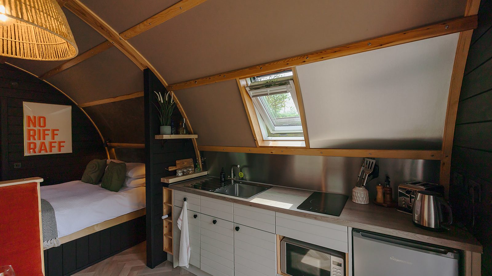 A cozy cabin interior with a small kitchenette, microwave, sink, and a skylight above. The bed is nestled beside a wall poster, all embraced by hewn wooden walls. It’s the perfect Yorkshire retreat.