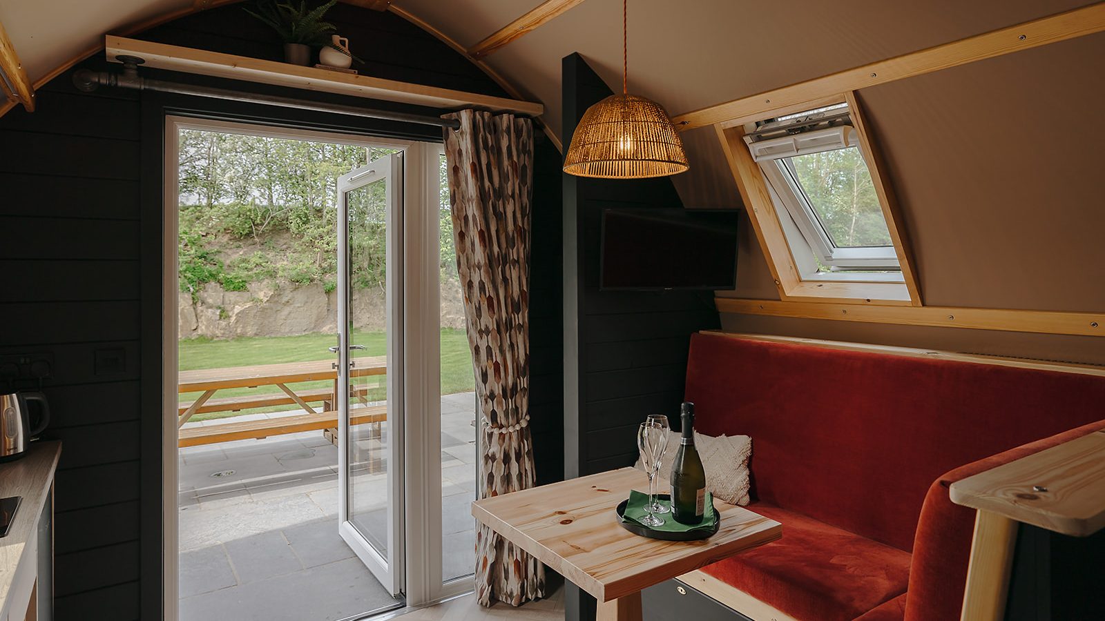 The interior of the cozy cabin features a hewn table, a red cushioned bench, and a pendant light. Open doors lead to a charming picnic area outside, reminiscent of the tranquil Yorkshire countryside.