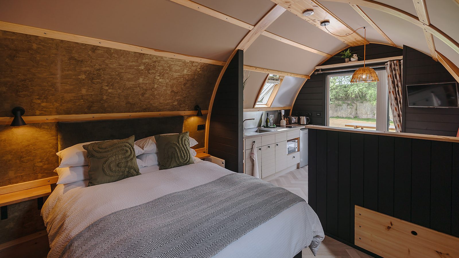 Cozy cabin interior featuring a neatly made bed, a hewn wood kitchenette, and large windows with a view of the Yorkshire outdoors.