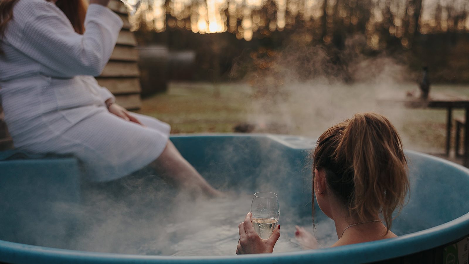 Two people unwind with drinks in a steaming outdoor hot tub at sunset, one seated on the edge, the other in the water, surrounded by hewn stone. The serene setting echoes the tranquil beauty of Yorkshire's landscapes.