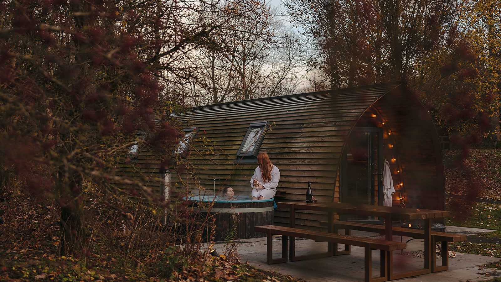 A hewn wooden cabin with a warm, lit interior stands serenely amidst Yorkshire trees. Outside, a person relaxes in a hot tub near a table and bench, enjoying the crisp fall evening.