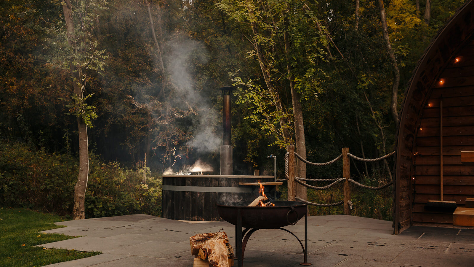 An outdoor scene in Yorkshire features a hewn fire pit with neatly stacked logs, next to a steaming wooden hot tub. The area is embraced by towering trees and a rustic wooden building, creating a serene natural retreat.