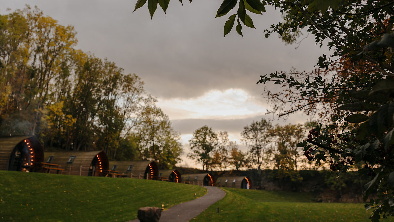 A path leads through a grassy area lined with pod-like cabins, surrounded by trees under a cloudy sky. The cabins feature handcrafted Yorkshire stone accents, adding a rustic charm to the serene landscape.