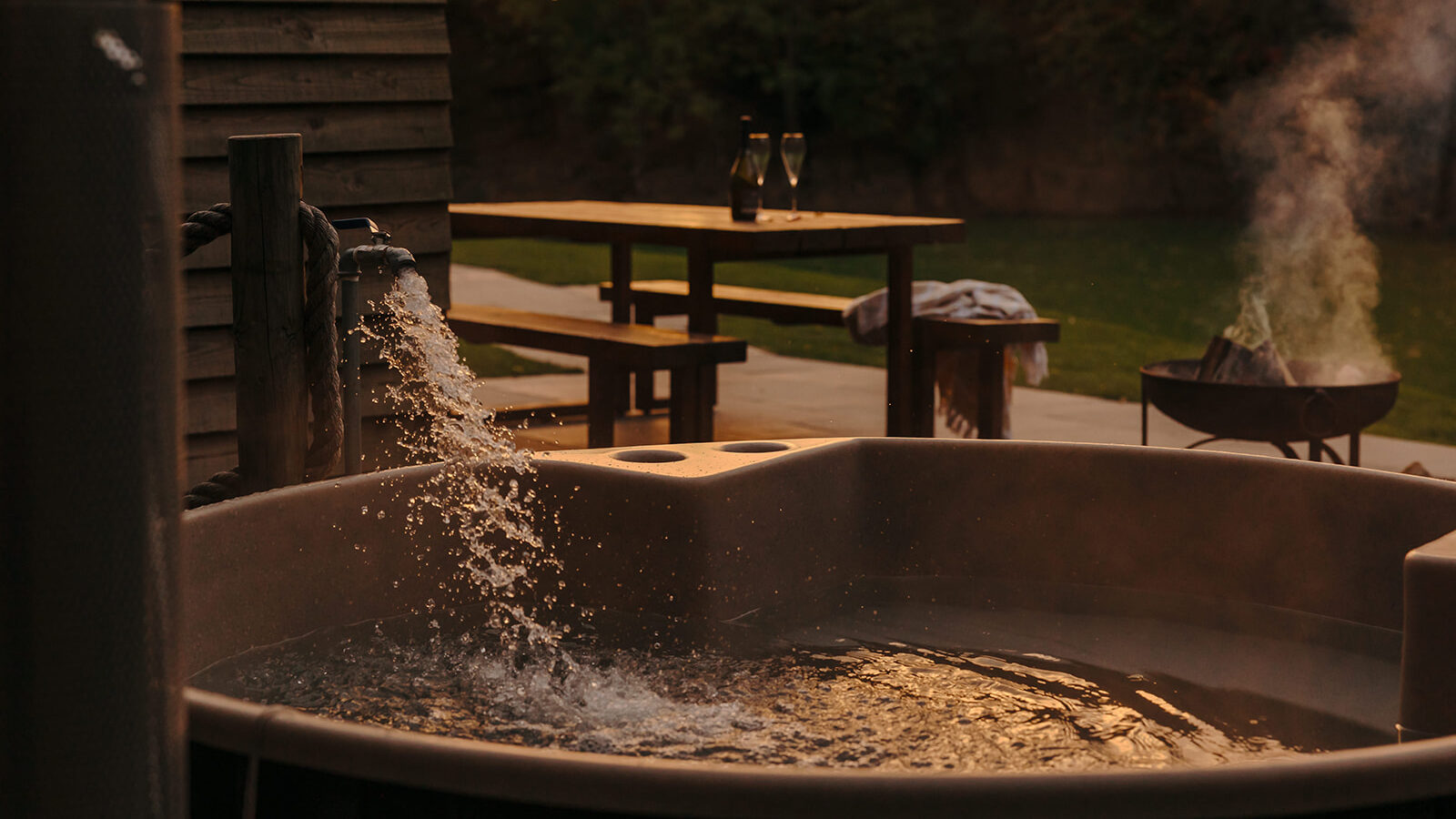 At sunset, a hot tub hewn from local materials fills with water beside an outdoor seating area and fire pit, offering a serene Yorkshire setting.