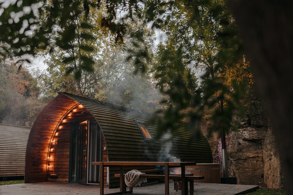 A cosy wooden cabin with a curved roof is nestled among lush trees, perfect for winter glamping in the UK. Warm lights glow from the cabin’s interior. Outside, there is a picnic table and a fire pit emitting smoke, creating a serene and inviting atmosphere in this natural setting. Hewn Yorkshire Glamping