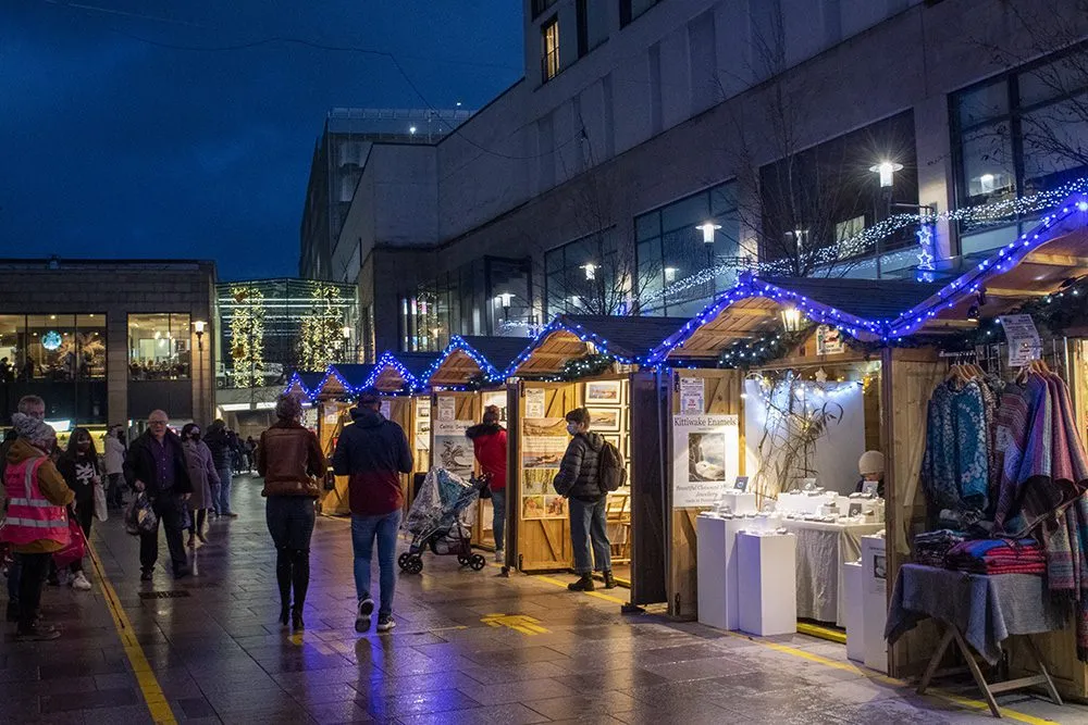 People stroll through one of the best Xmas markets in the UK at night, with illuminated stalls and blue string lights twinkling overhead.