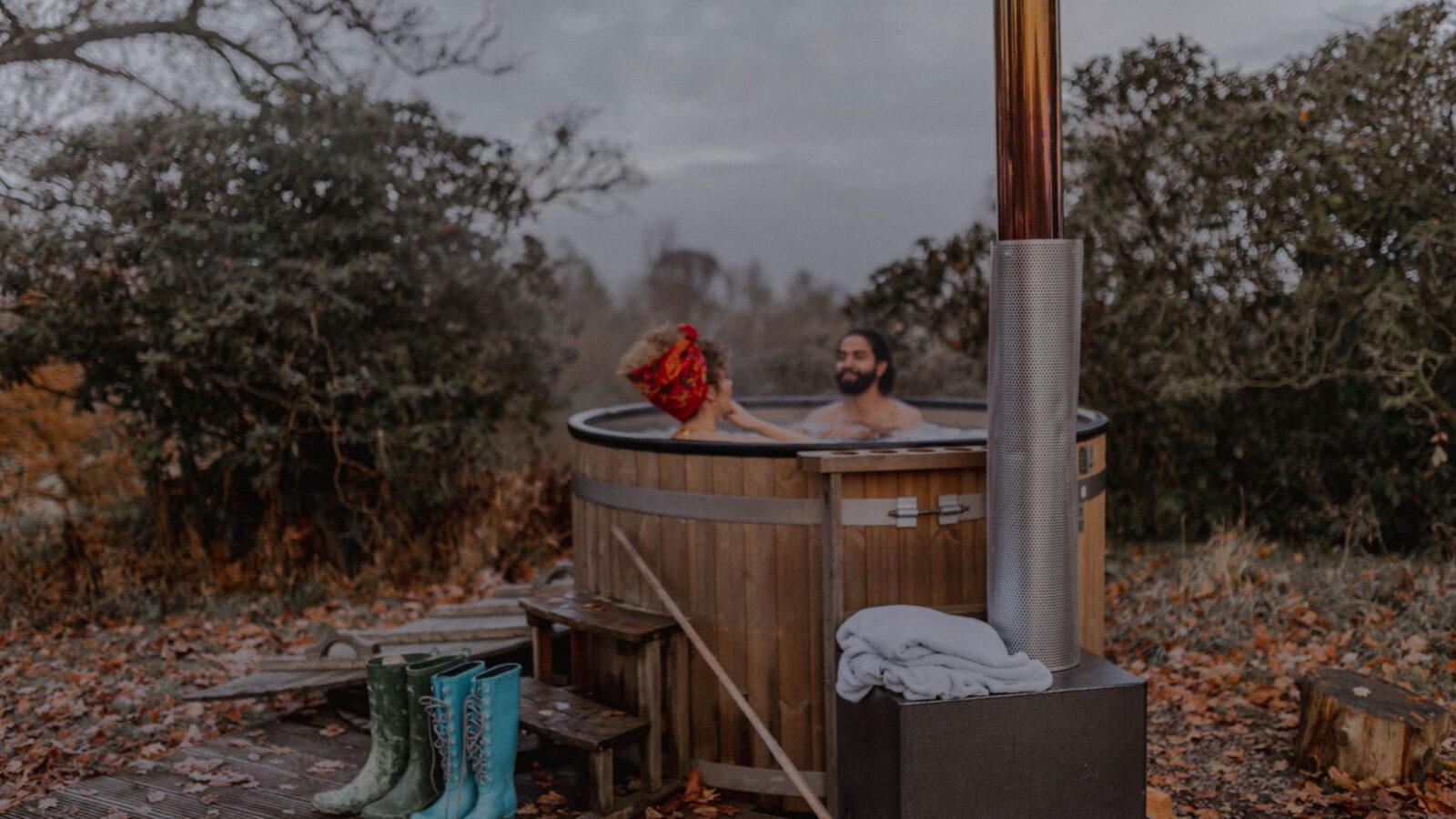 A person relaxes in a wooden hot tub outdoors at the Netherby Treehouse, surrounded by autumn leaves, with boots and a towel on the deck nearby.