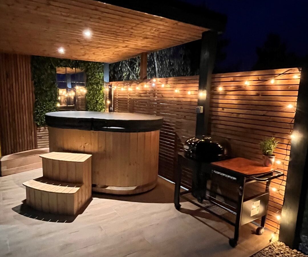 Nestled on a deck under a canopy of warm string lights, the wooden hot tub at New Farm Glamping creates a perfect retreat. Nearby, a small grill stands ready for your culinary adventures, enhancing this serene night with the true essence of glamping. 