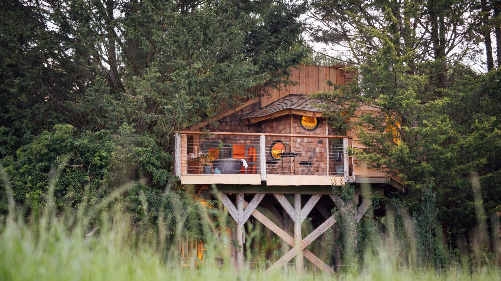 Nestled in an orchard, this treehouse boasts a wooden deck surrounded by trees and tall grass, featuring circular windows and a cozy design inspired by Fenny Castle's charm.