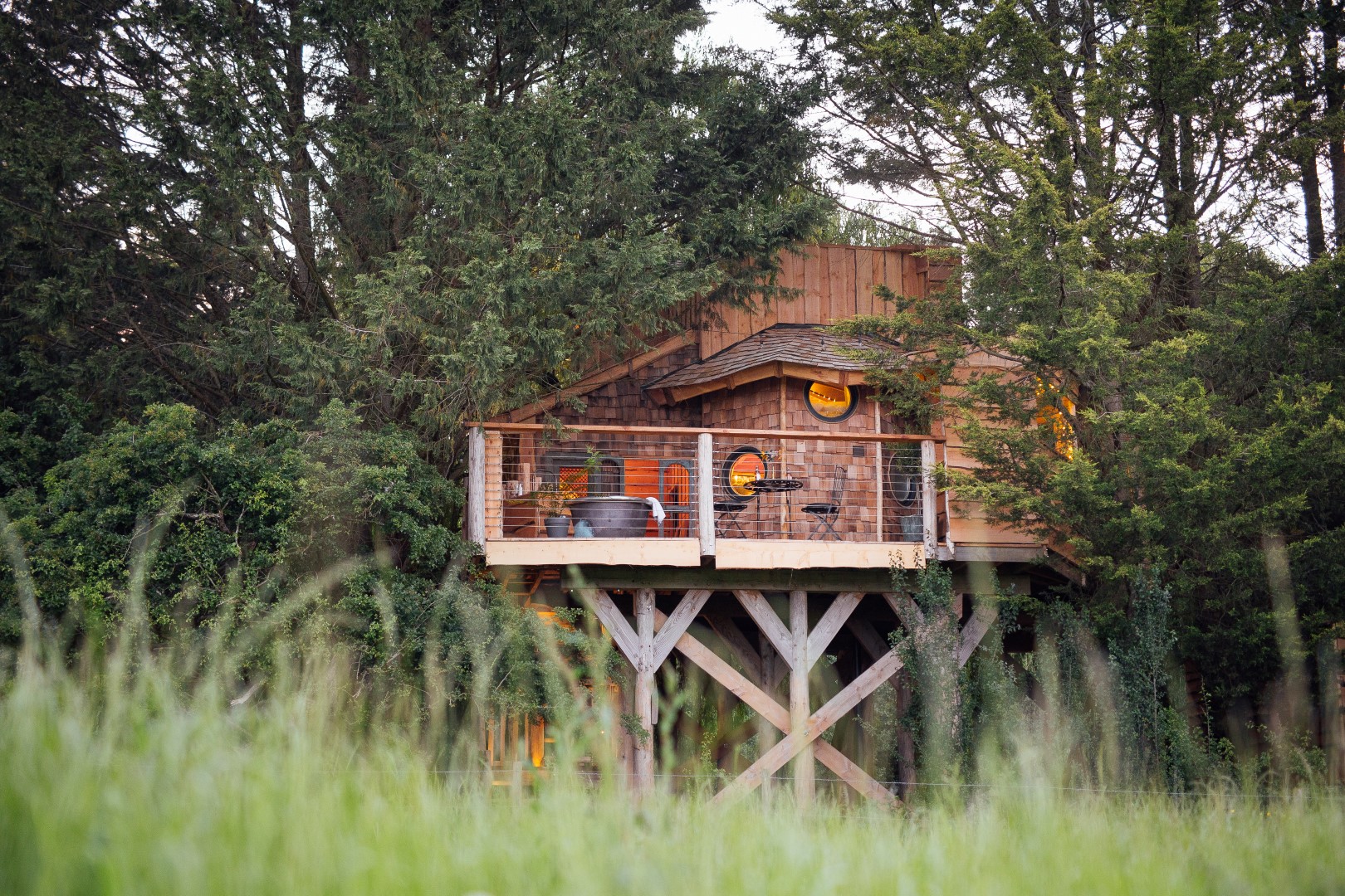 Nestled in an orchard, this treehouse boasts a wooden deck surrounded by trees and tall grass, featuring circular windows and a cozy design inspired by Fenny Castle's charm.