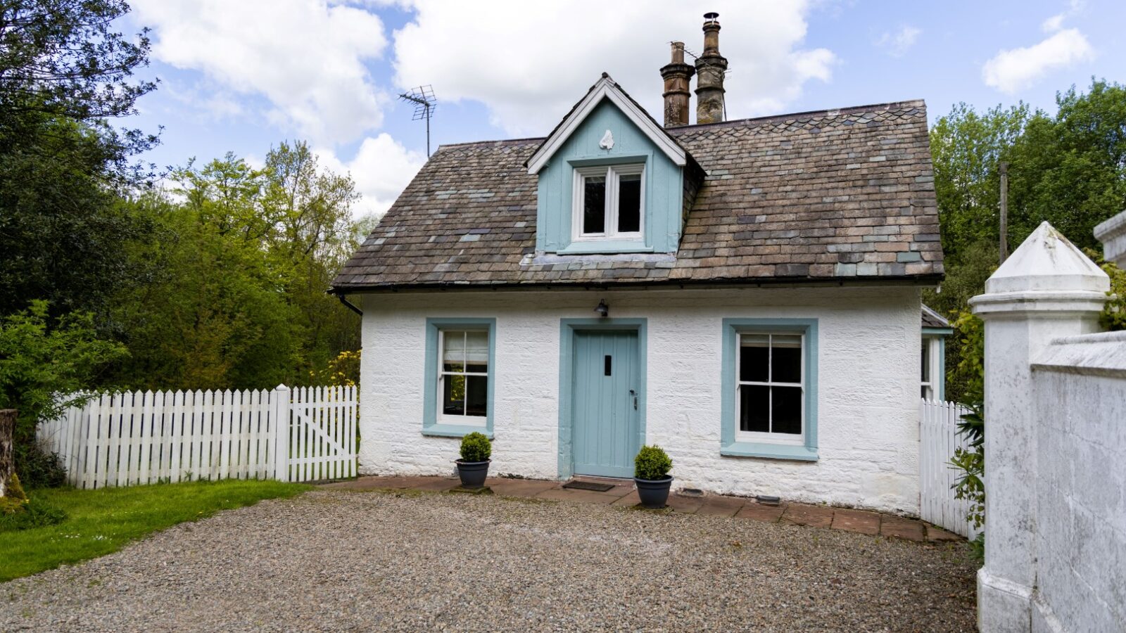 A cozy, white cottage with blue trim and a gray shingled roof, nestled among trees and a white picket fence, offers the charm of a Top Lodge accommodation.