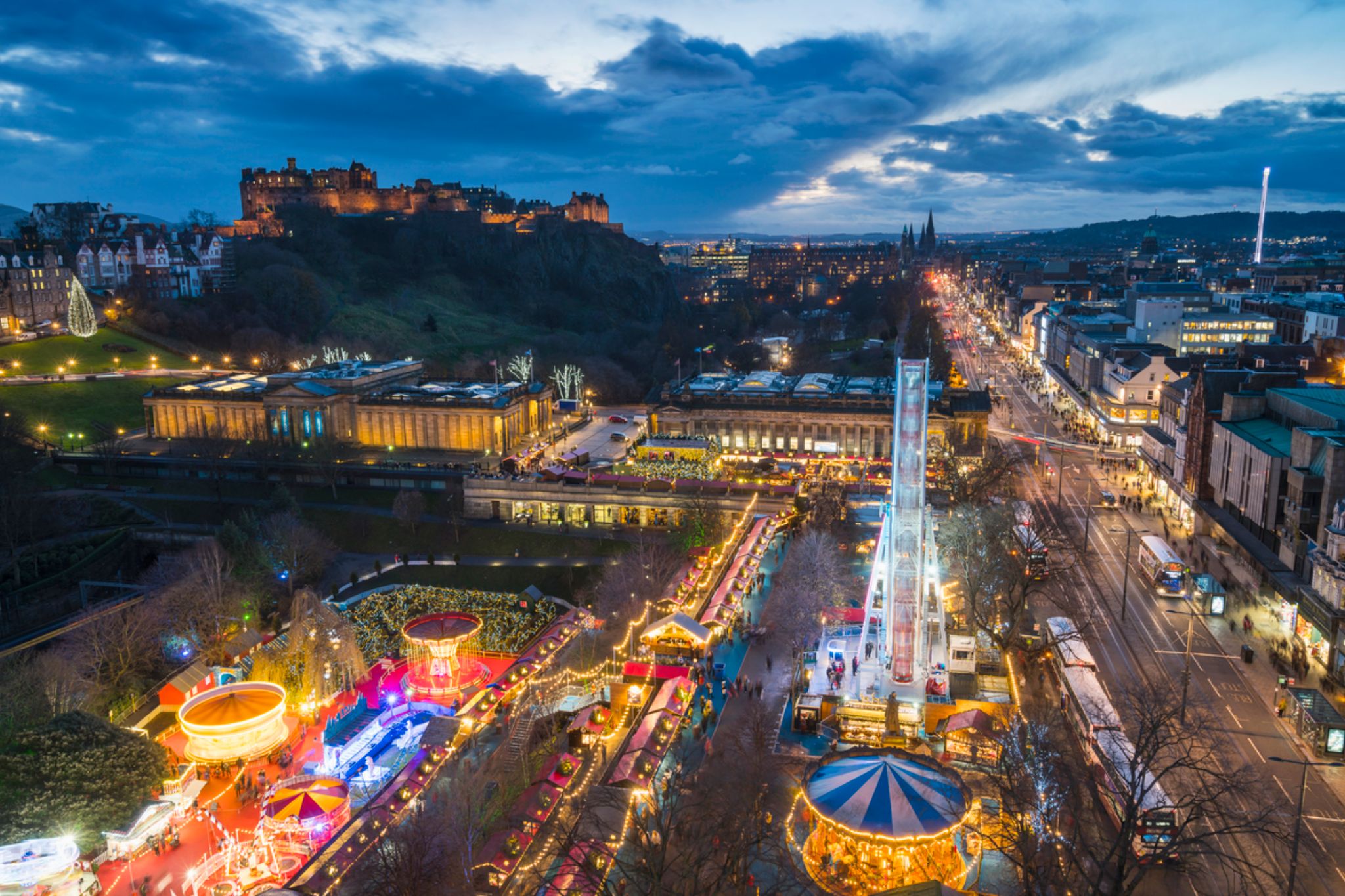 An aerial view of the cityscape at dusk reveals a lit-up fair with a dazzling Ferris wheel and a castle perched on a hill, reminiscent of the best Xmas markets in the UK, where magic dances in the air.