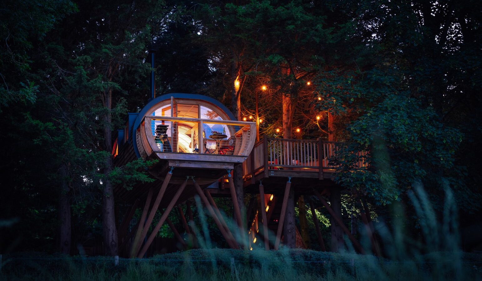 Nestled in an orchard, a treehouse with circular windows and string lights stands elevated on stilts at dusk, offering enchanting views reminiscent of Fenny Castle's storied landscapes.