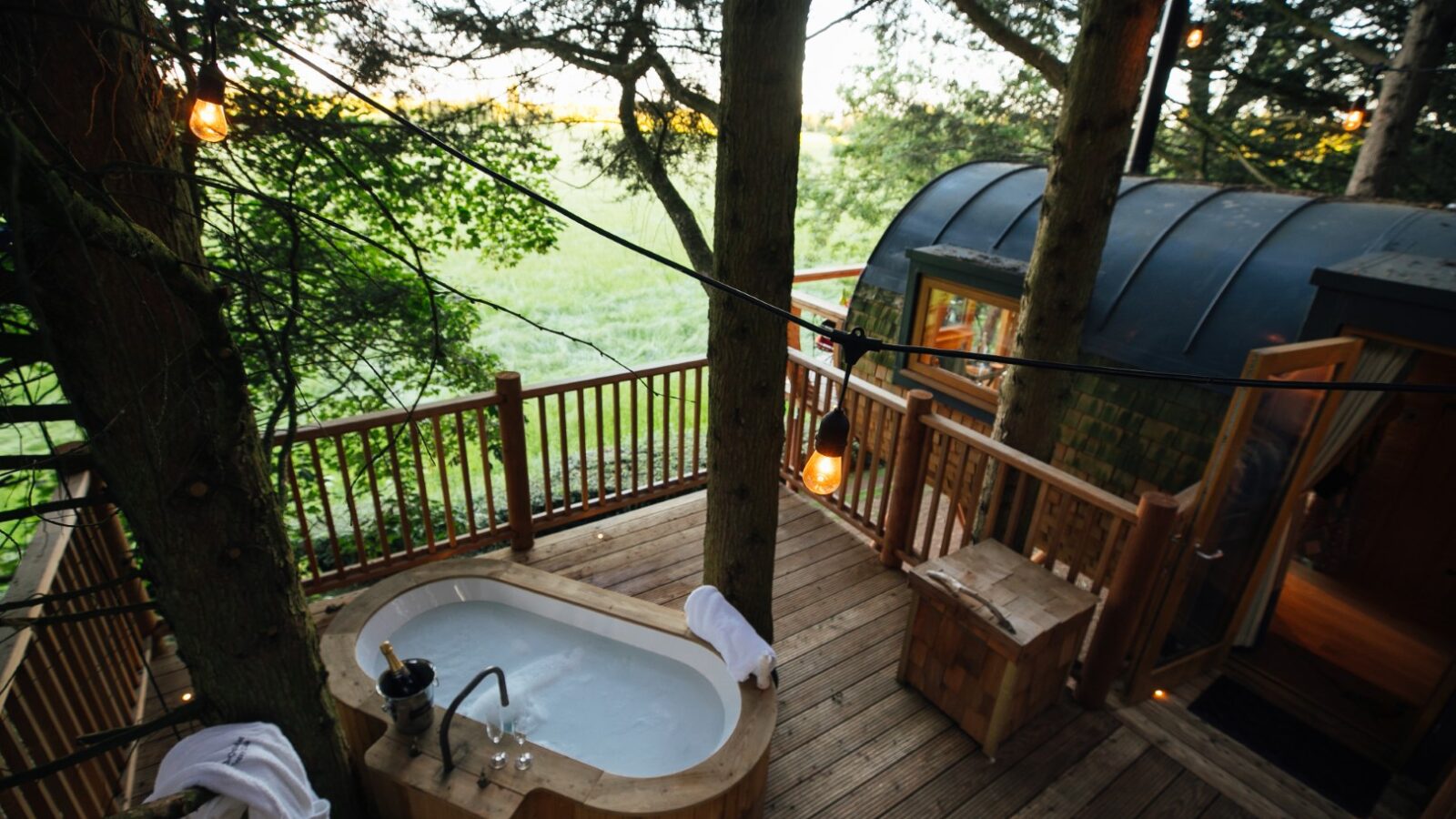 A wooden deck with a freestanding bathtub, nestled amidst trees and glowing string lights near a small cabin with an open door, offers a serene escape at Fenny Castle.