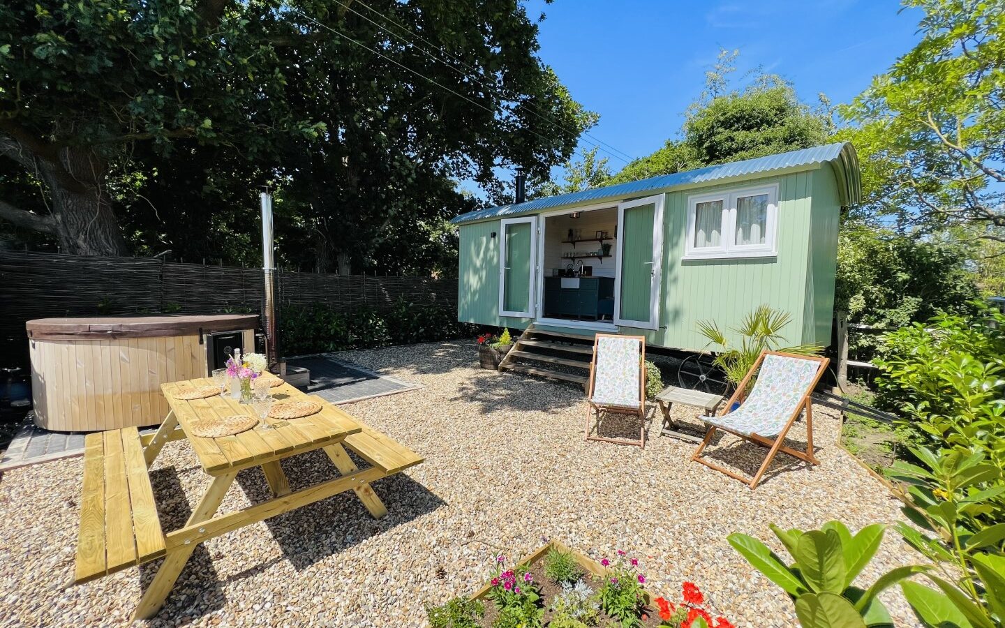 The charming green hut features a gravel patio with a picnic table, hot tub, two deck chairs, and a lush garden, all set in a sunny outdoor haven.