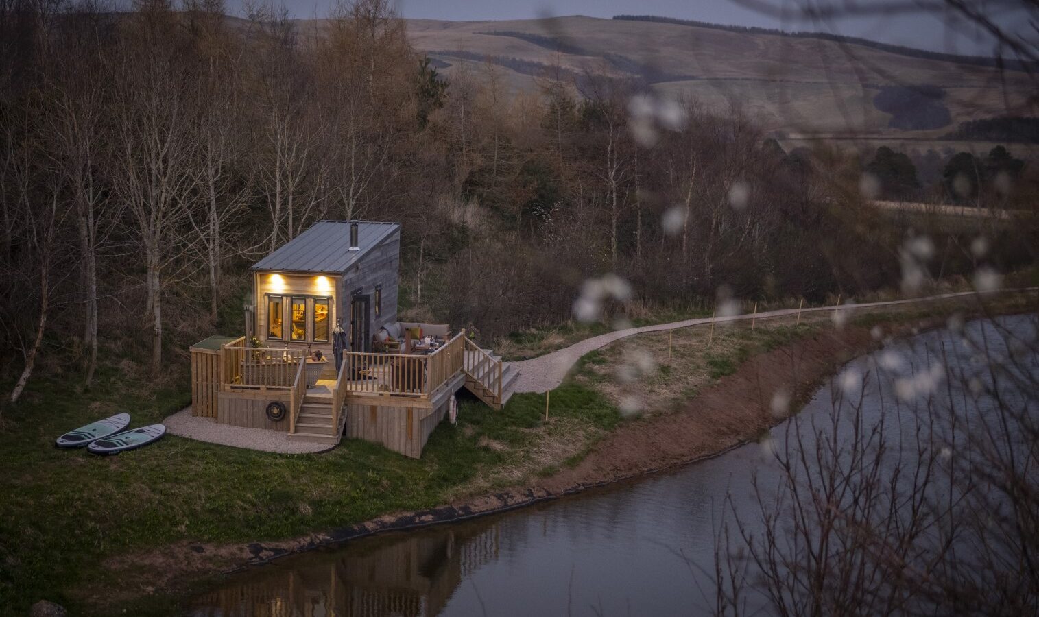 A tiny home with a warm, lit interior rests by a lake, bordered by bare trees and hills under a dusky sky. Two paddleboards are nearby.