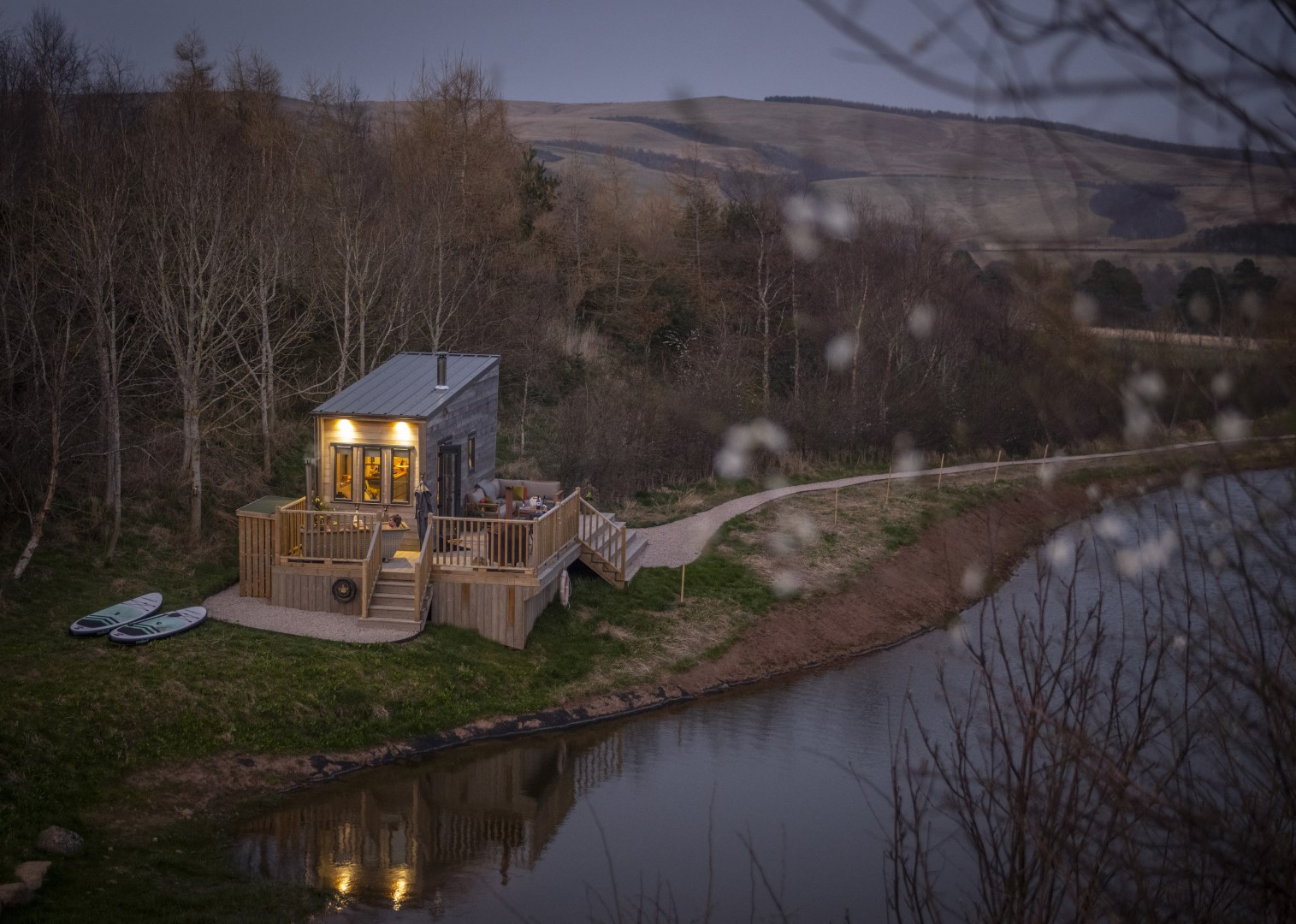 A tiny home with a warm, lit interior rests by a lake, bordered by bare trees and hills under a dusky sky. Two paddleboards are nearby.