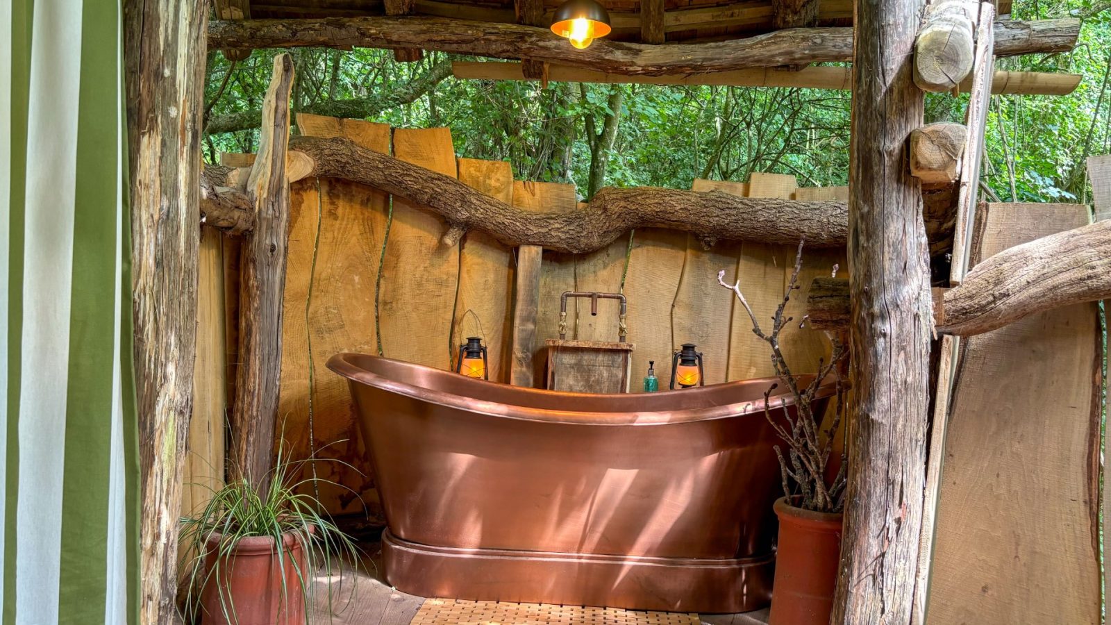 A rustic outdoor bathroom nestled in One Acre Wood features a copper bathtub surrounded by wooden walls and leafy green plants. Dim lighting enhances the natural, cozy atmosphere, while earthy decor is enriched by natural wooden beams and elements.