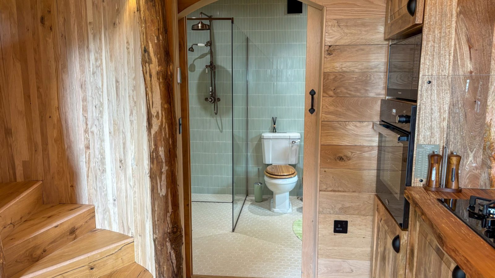 Nestled in One Acre Wood, a small wooden cabin bathroom boasts a shower with glass walls, a white toilet with a wooden seat, and light green tiles. The cabin's rustic charm comes alive in its wooden walls. Stairs lead up to the left side of the image.