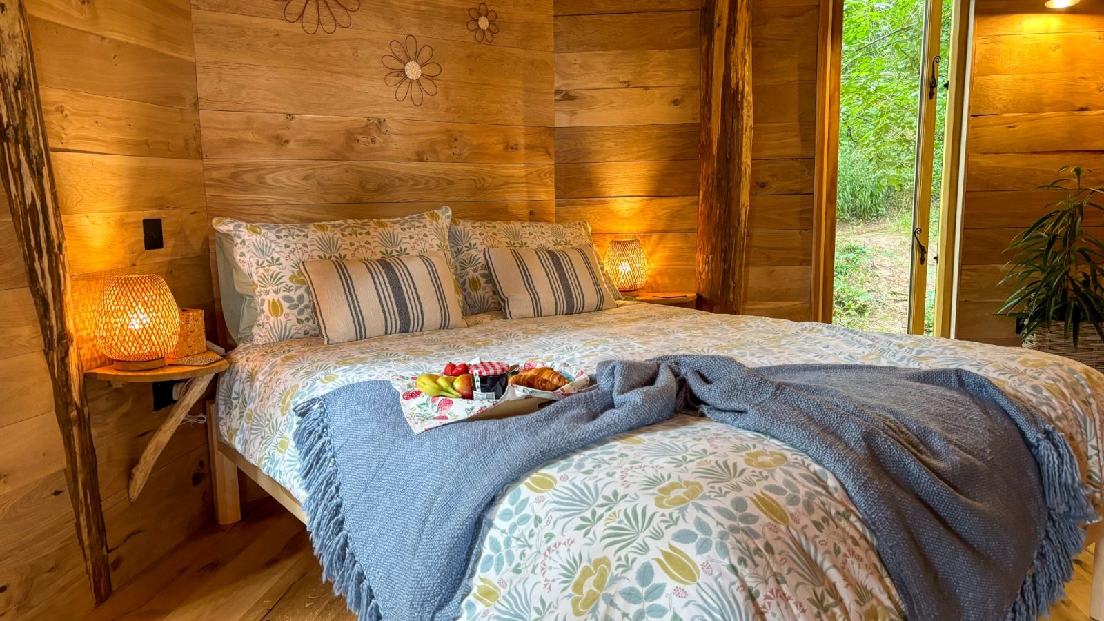 Cozy bedroom with a wooden interior inspired by One Acre Wood, featuring a floral-patterned bedspread and decorative pillows. Two wooden lamps illuminate the room. A tray with fruit and croissants sits on the bed. A large window reveals a view of lush greenery outside.