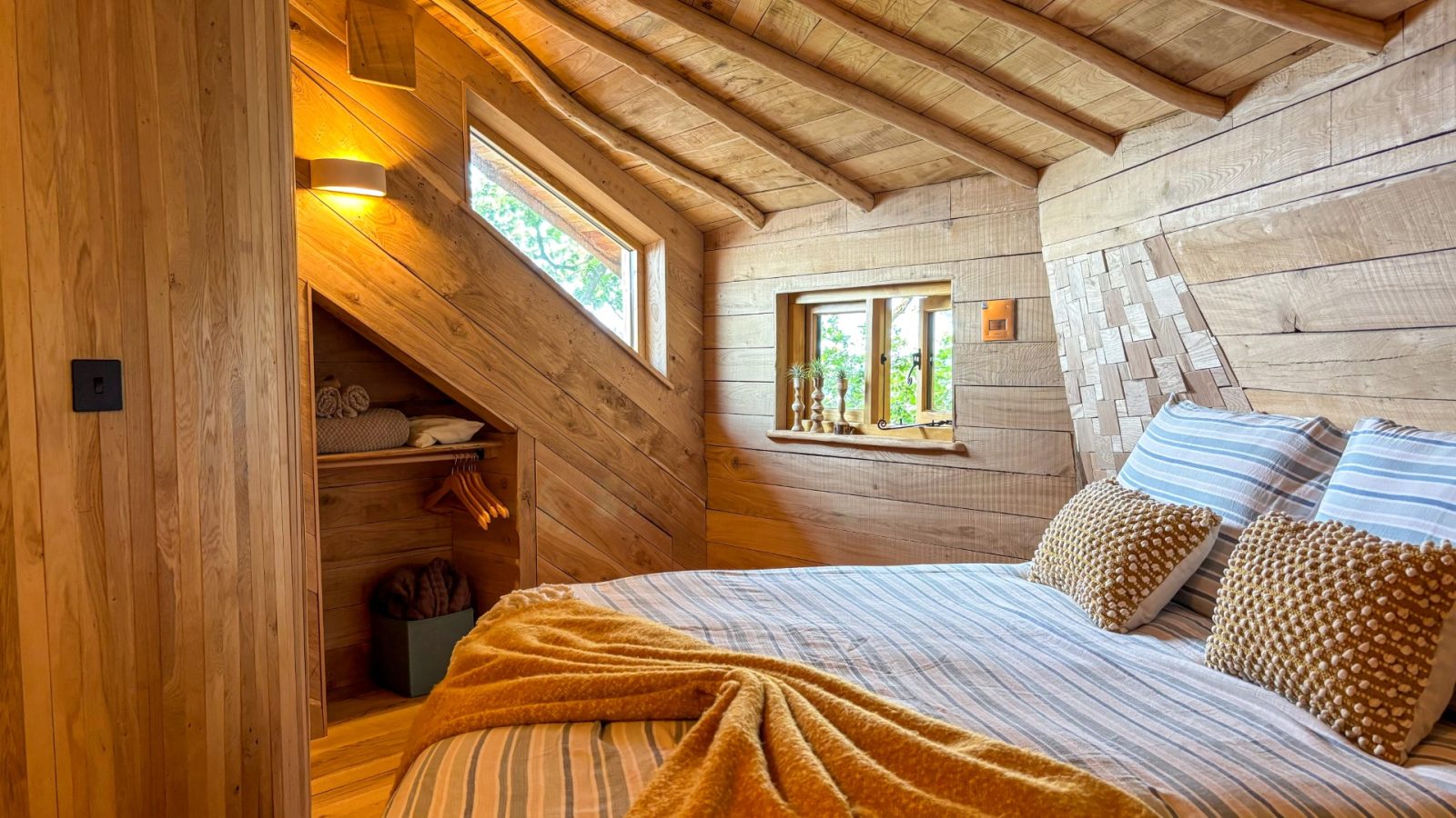 A cozy wooden bedroom in One Acre Wood features a sloped ceiling and a bed adorned with striped linens and mustard-colored pillows. A warm throw is draped over the bed, while shelves for storage and a small window complete the rustic, inviting atmosphere.