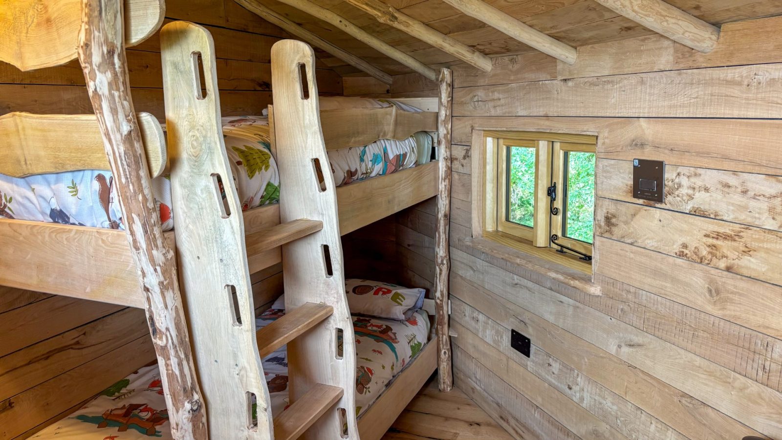 A cozy rustic cabin room with wooden walls and ceiling, nestled in the heart of One Acre Wood. It features a bunk bed with colorful bedding, where a small window lets in natural light. A sturdy wood ladder leads to the top bunk, completing this charming woodland retreat.